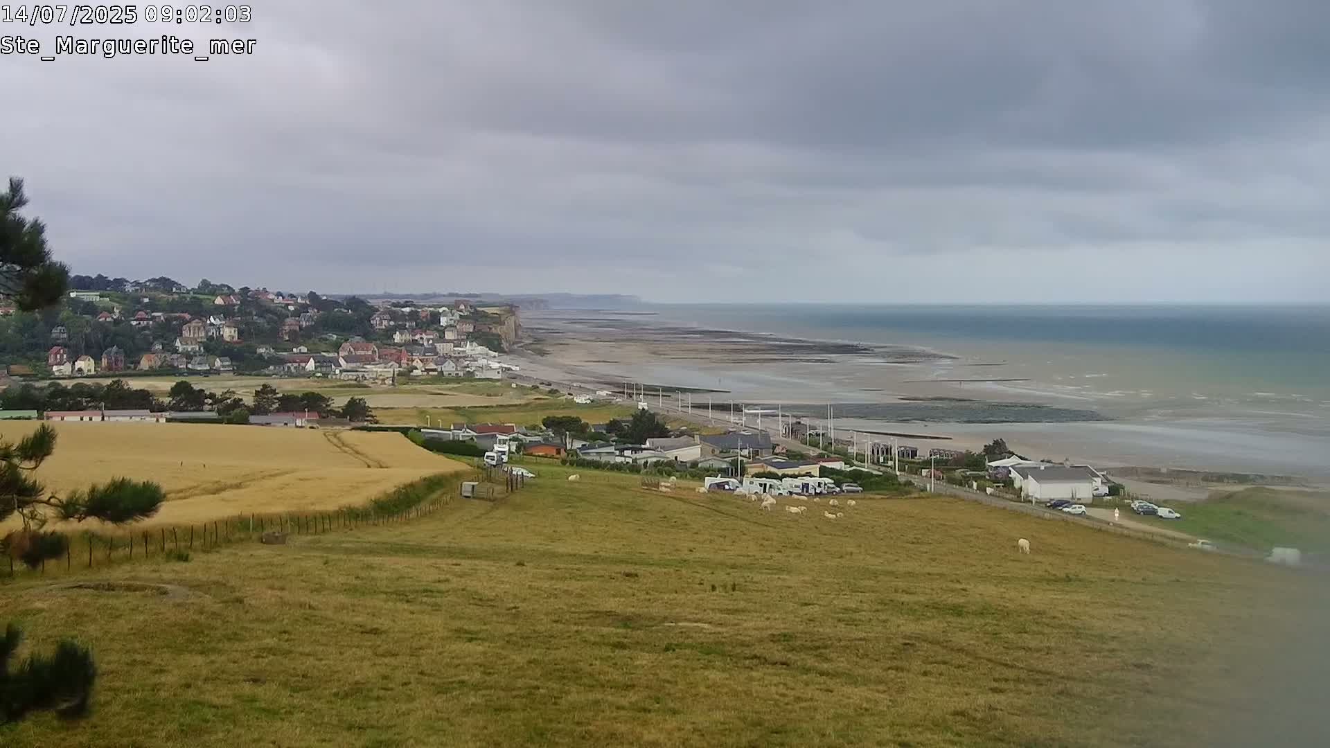 A coastal town is visible from a high vantage point on an overcast day, overlooking a wide expanse of sandy beach and calm sea.