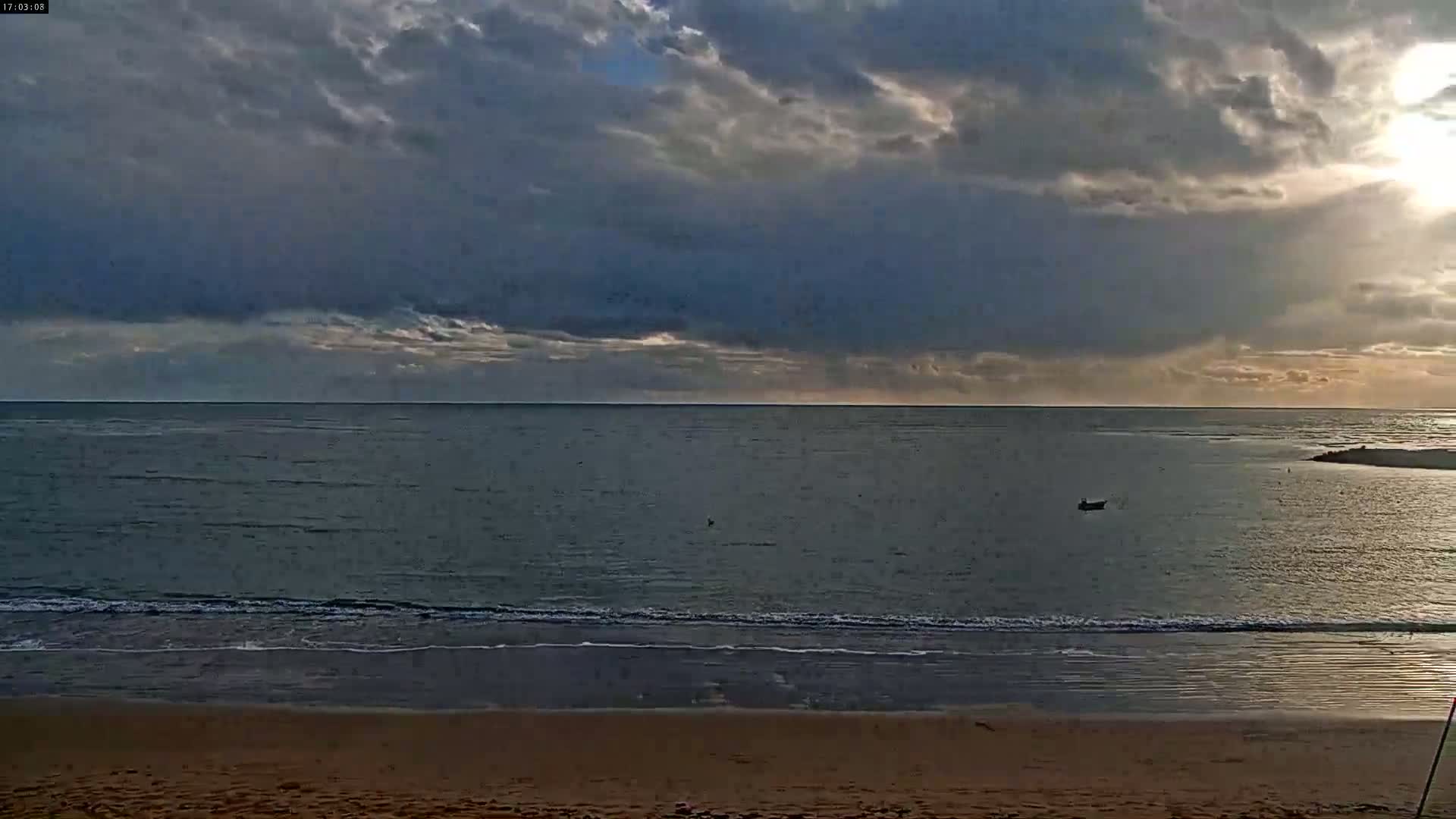 A sandy beach meets a calm sea under a partly cloudy sky, with a single small boat visible on the water.