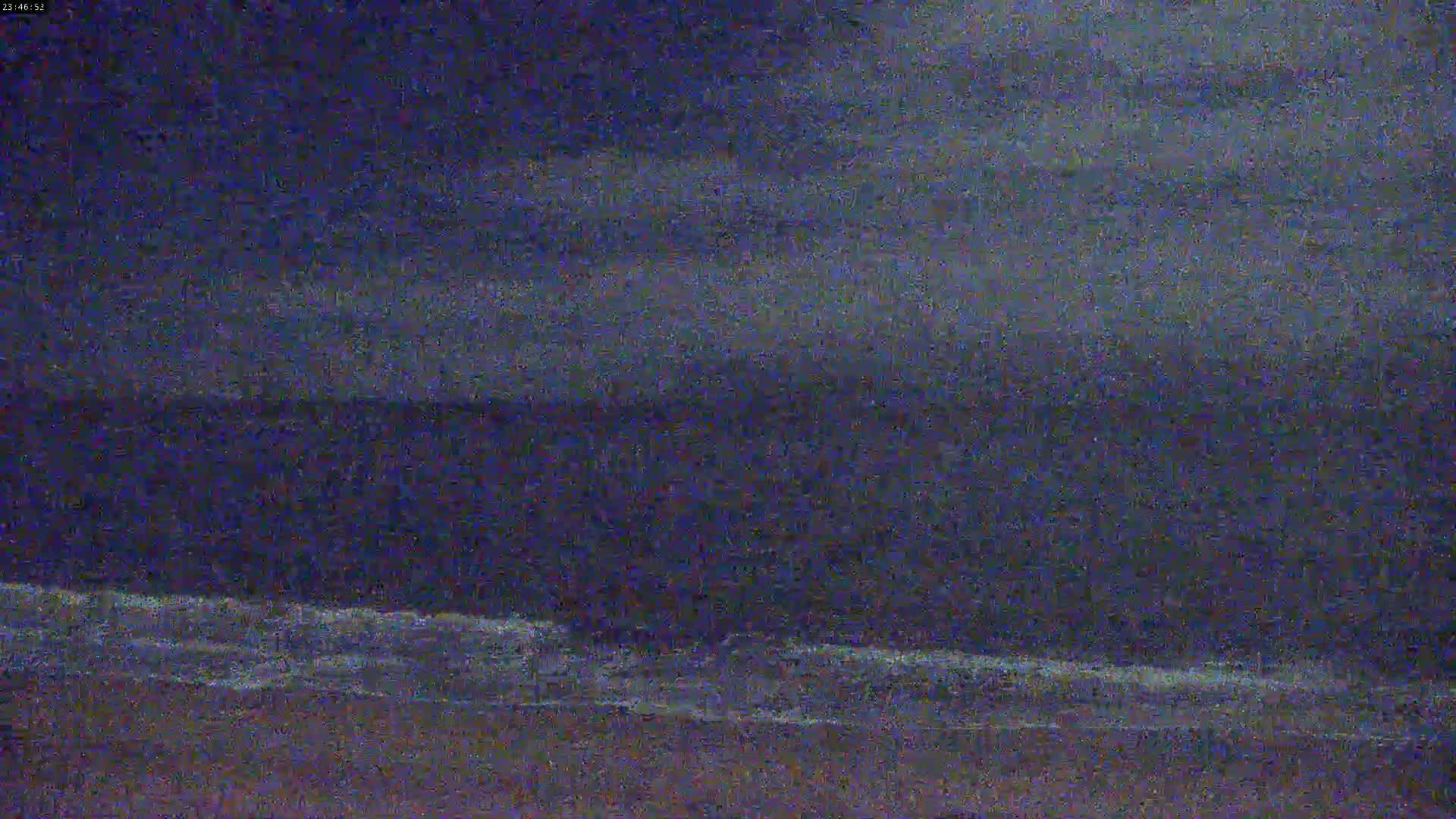A dark, nighttime beach scene shows a calm ocean with small waves lapping the shore under a cloudy sky.