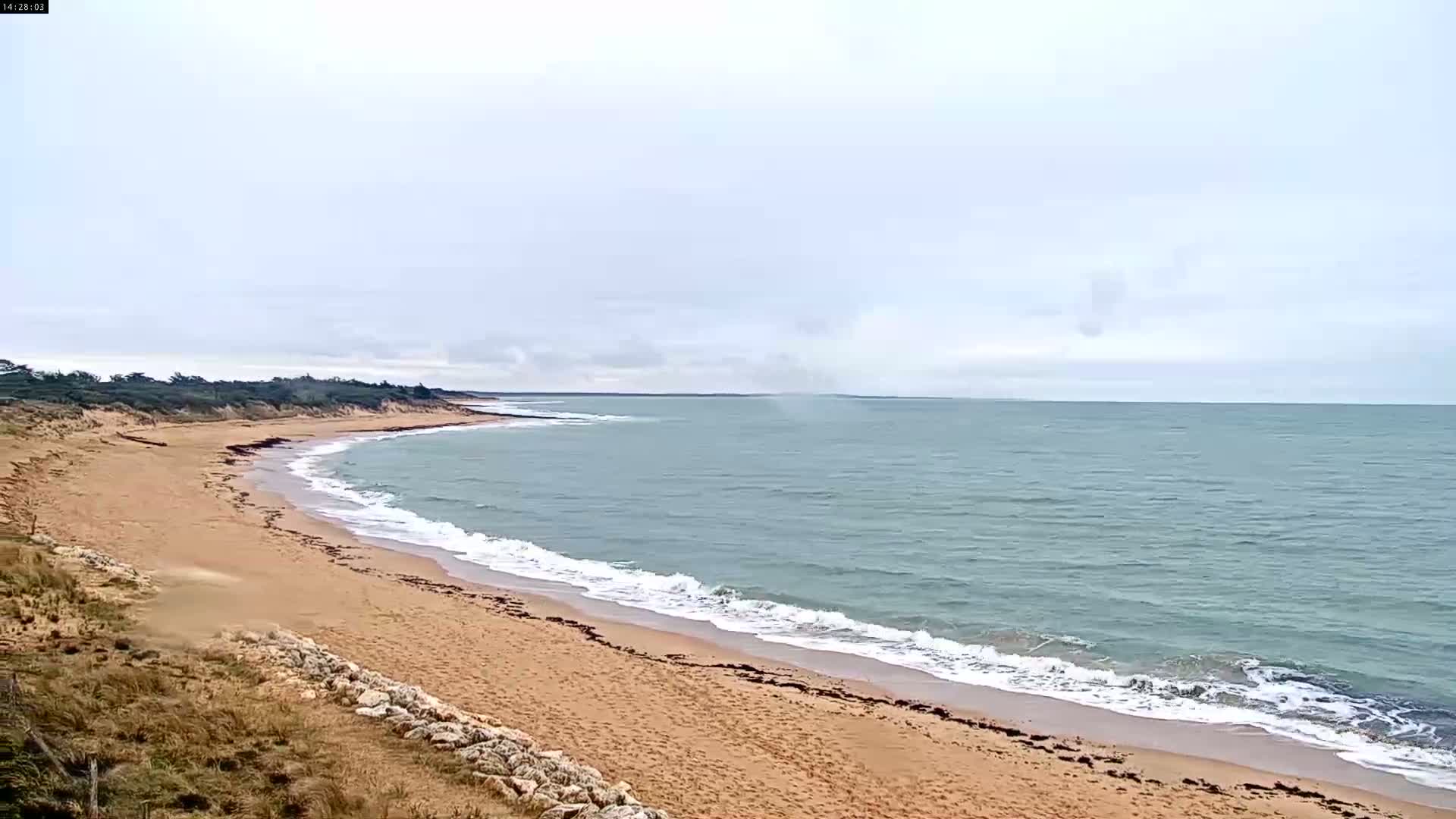 A sandy beach curves gently along a calm, light-blue ocean under a mostly cloudy sky.