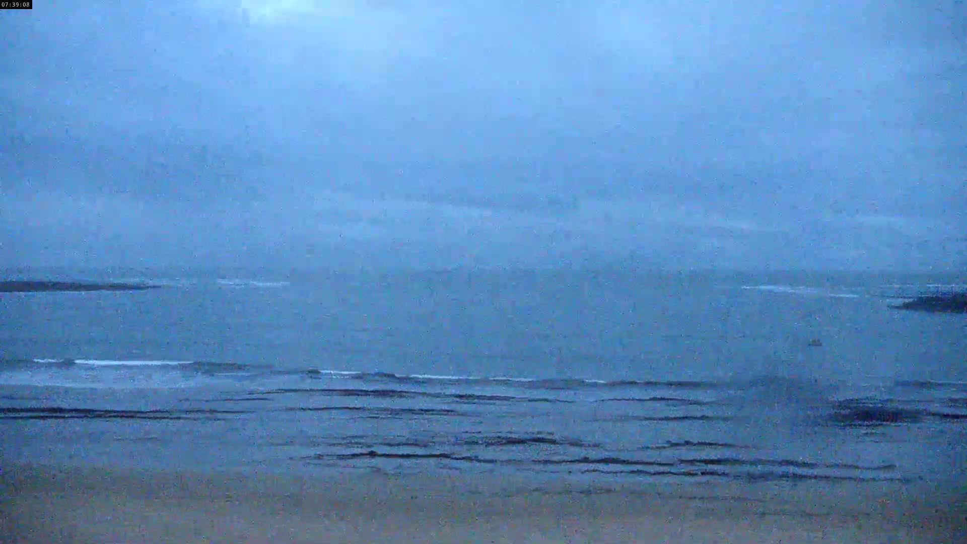 A mostly cloudy, overcast sky hangs over a calm ocean with small waves lapping a sandy shore.