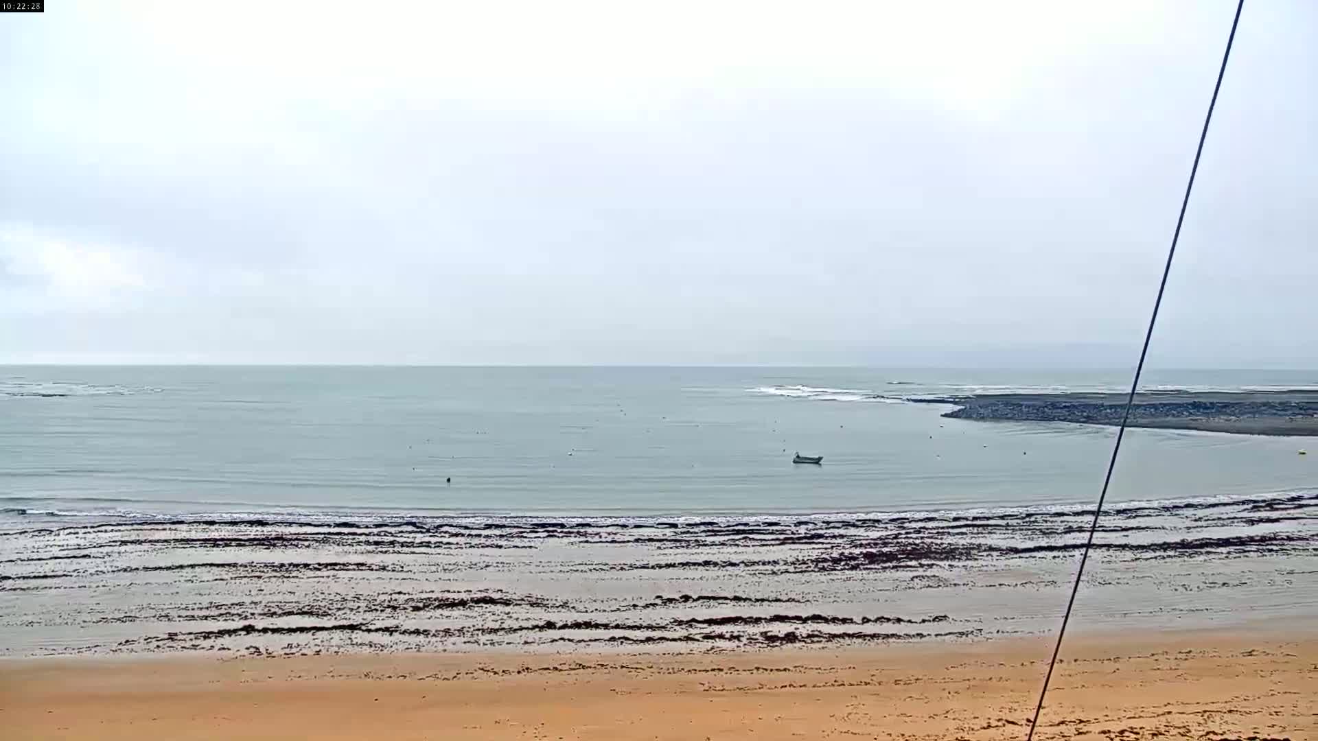 A calm, overcast day shows a sandy beach meeting a gently lapping ocean with a small boat visible in the distance.