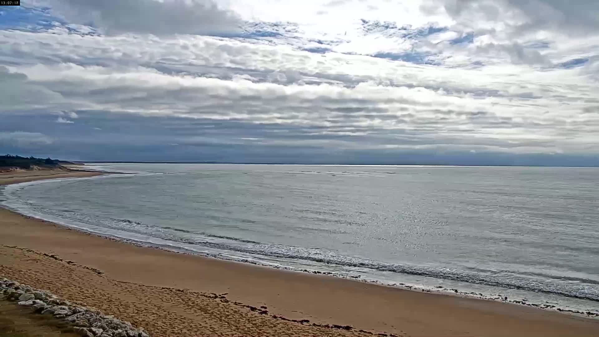 A sandy beach curves gently into a calm sea under a mostly cloudy sky.