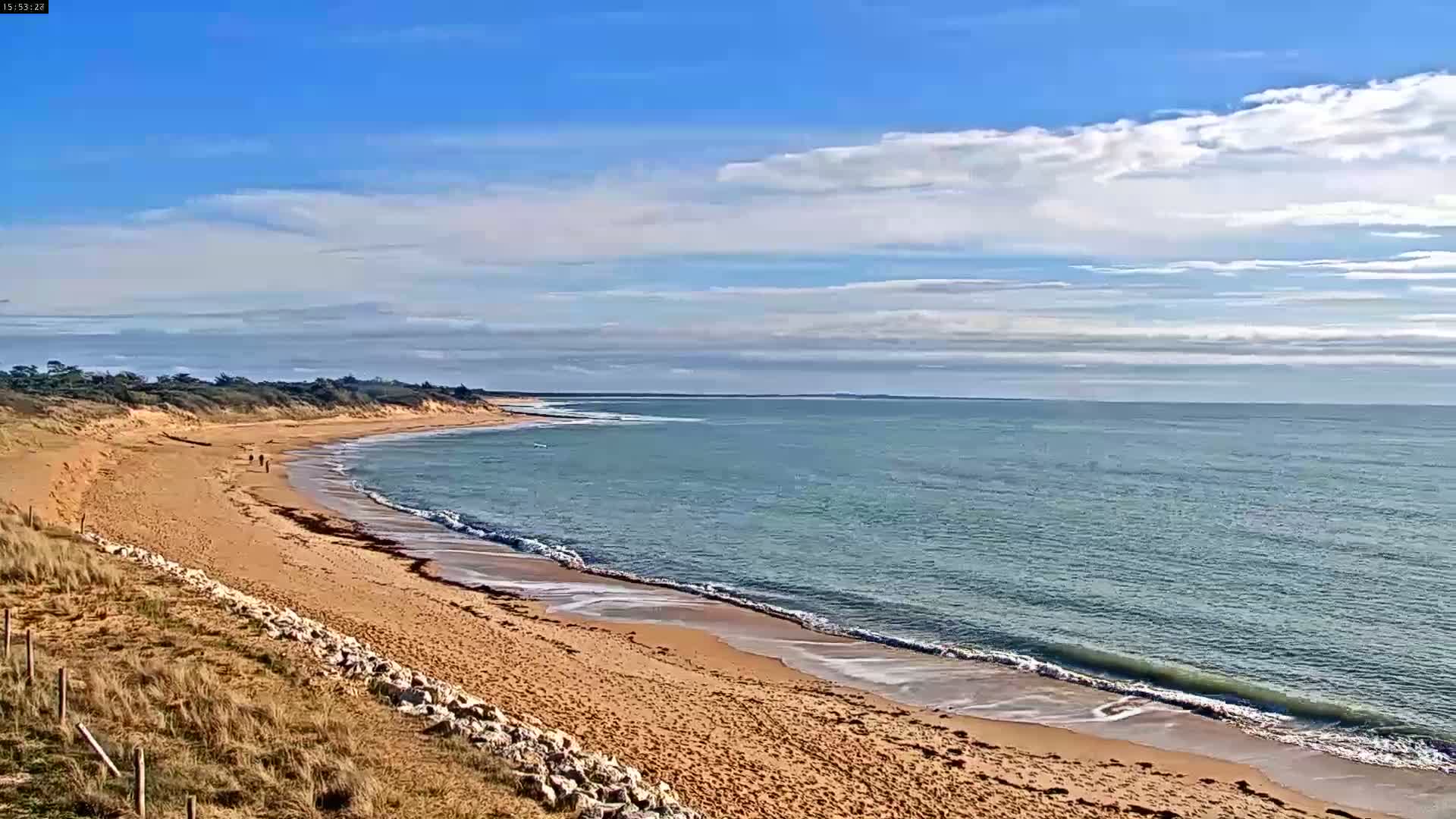 A sandy beach curves gently along a calm ocean under a mostly sunny sky with scattered clouds.