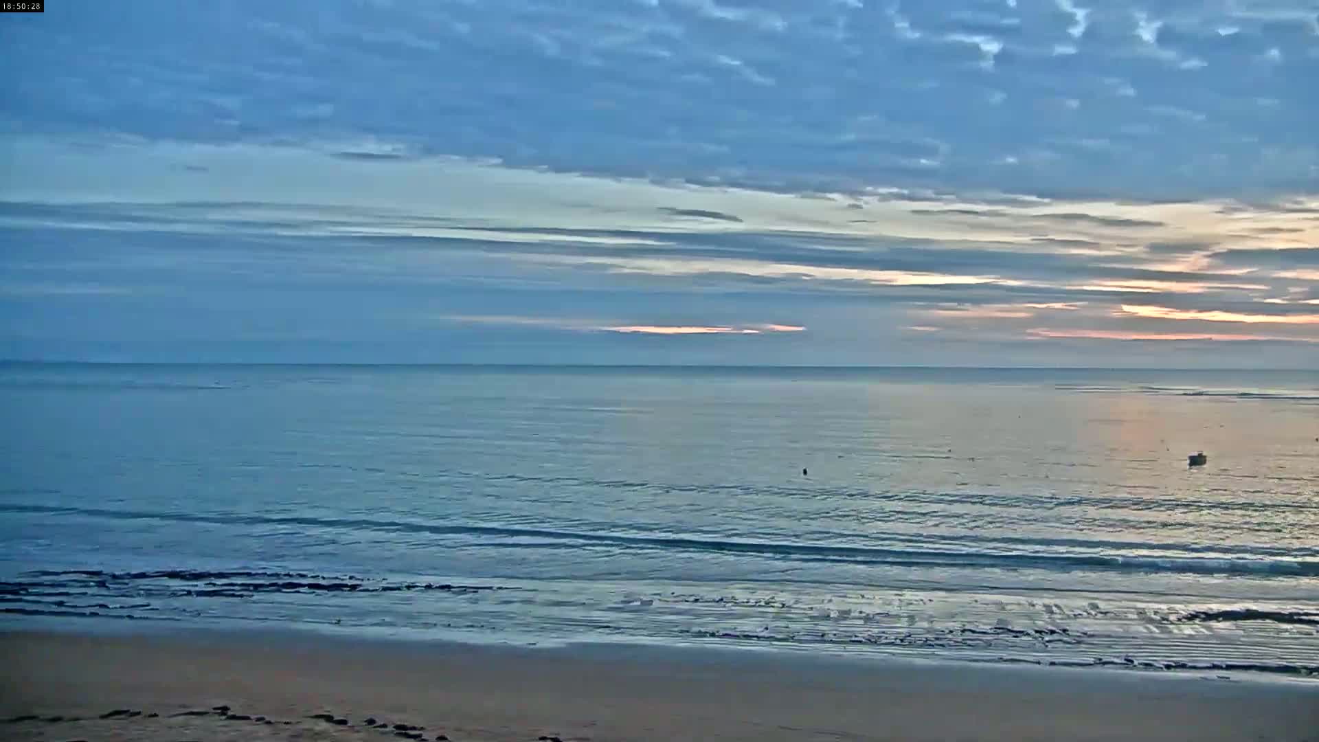 A sandy beach meets a calm ocean under a mostly cloudy, pale blue sky with hints of sunrise.