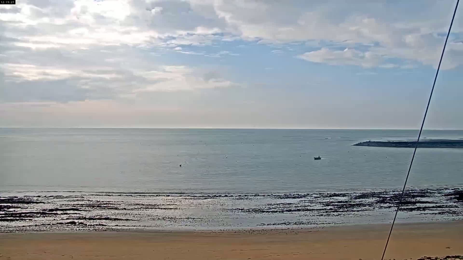 A mostly sunny day shows a calm sea with a sandy beach and a small boat in the distance.
