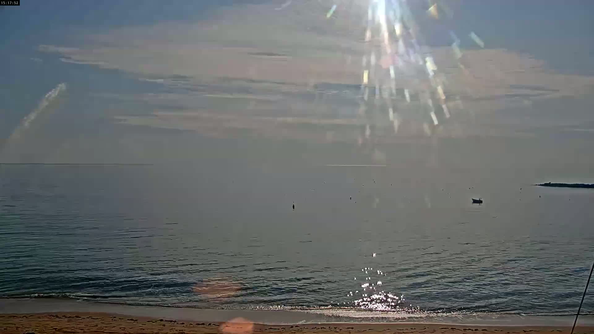 A sandy beach meets a calm sea under a mostly sunny sky with a few clouds and bright sun rays.