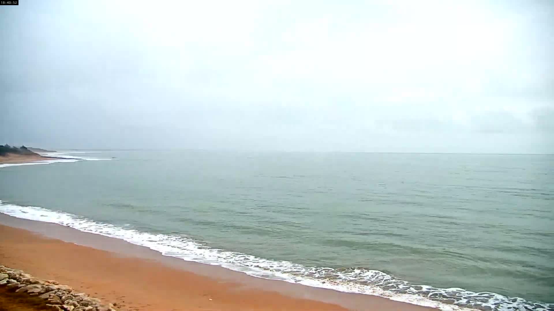 A sandy beach with gently lapping waves meets a calm, gray-green sea under an overcast sky.