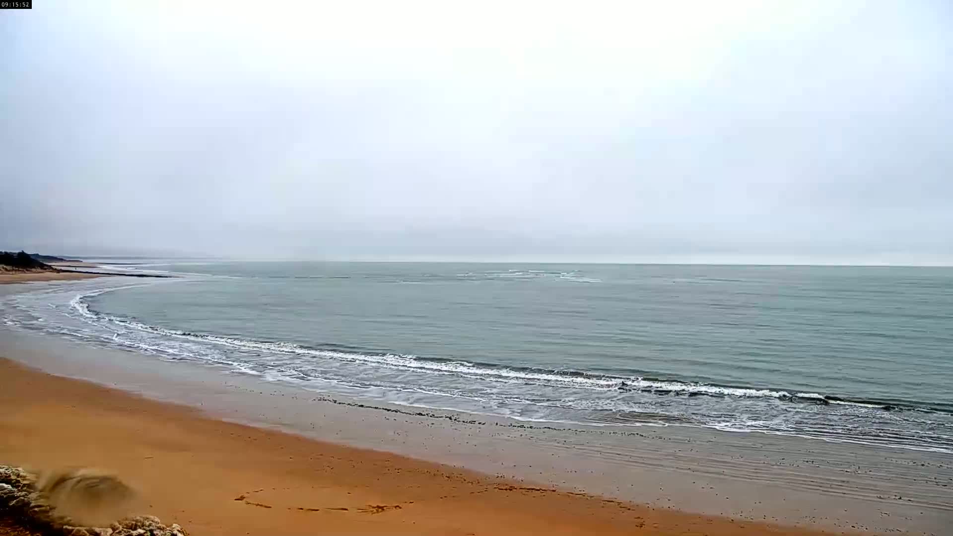 A sandy beach with gently rolling waves meets a calm, grey sea under an overcast sky.