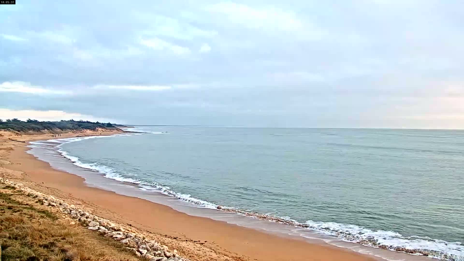 A long sandy beach curves gently alongside a calm, light-blue ocean under a mostly cloudy sky.
