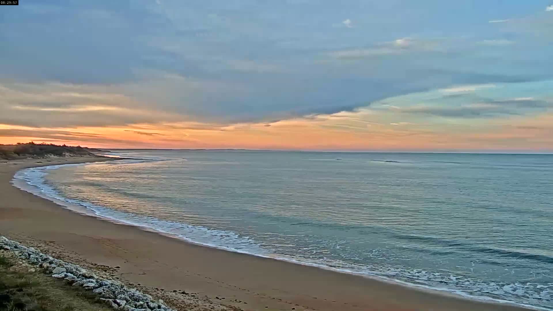 A sandy beach curves gently into a calm sea under a pastel sunset sky.