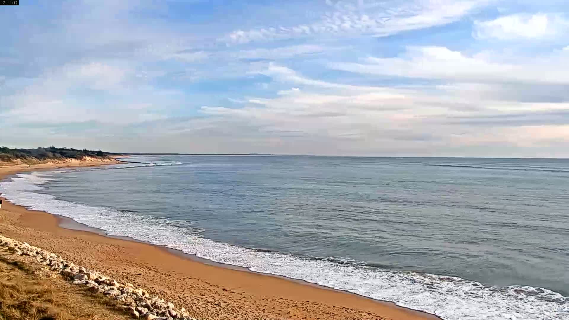 A sandy beach with gently rolling waves under a mostly clear, bright sky meets a calm ocean.
