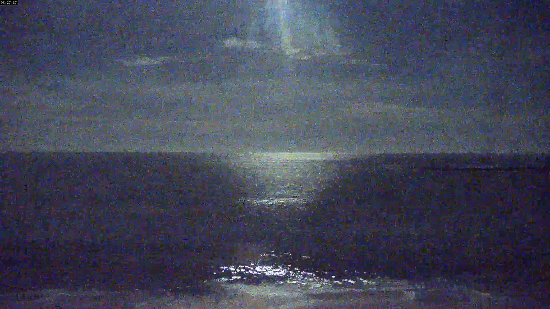 A dark, nighttime view of a calm body of water reflecting a bright light source in the sky under a mostly cloudy sky.