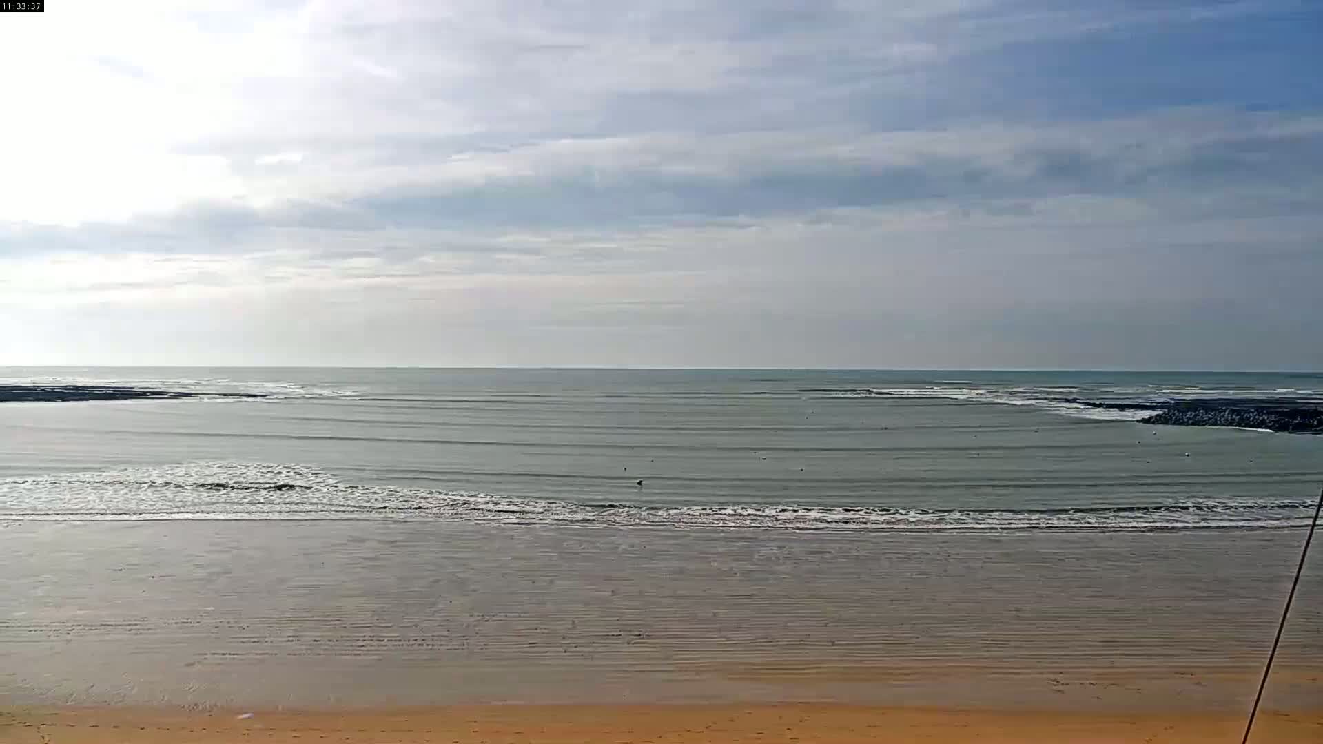 A sandy beach with calm, gray-green ocean waves under a mostly cloudy sky.