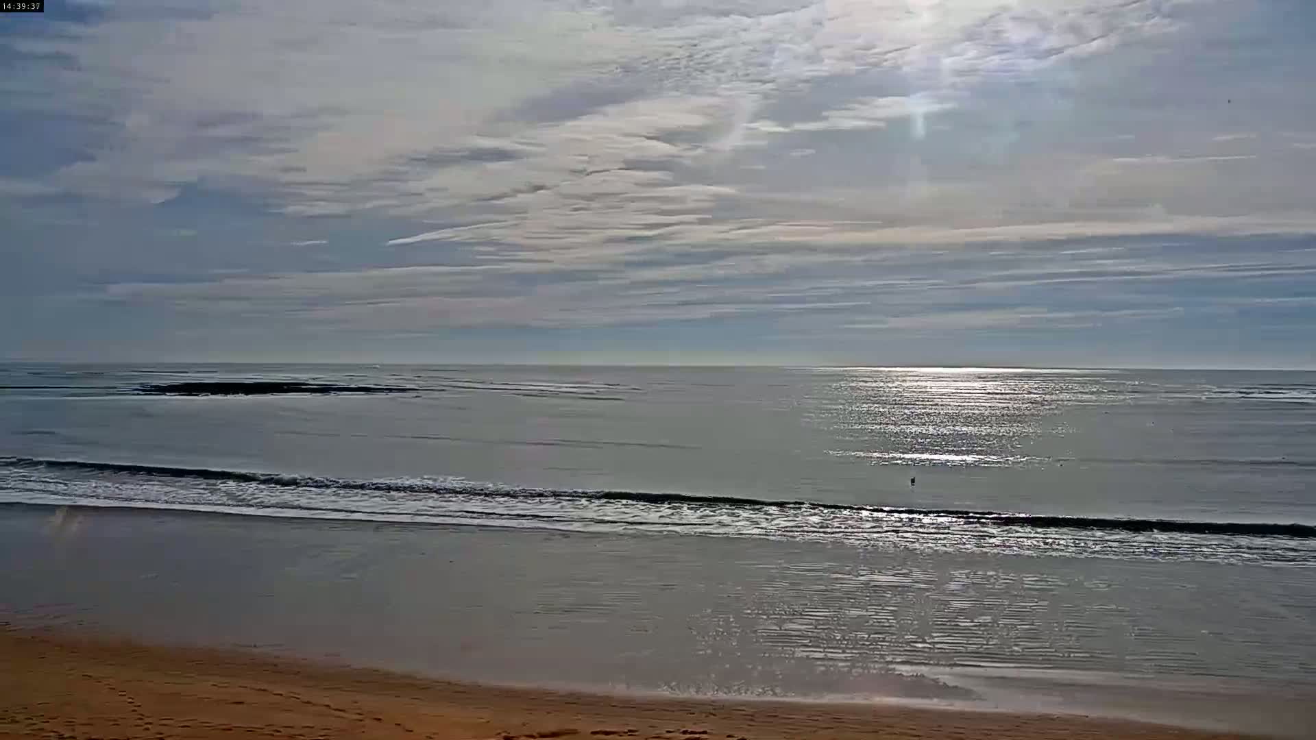 A mostly sunny day shows a calm ocean with small waves lapping a sandy beach under a lightly clouded sky.