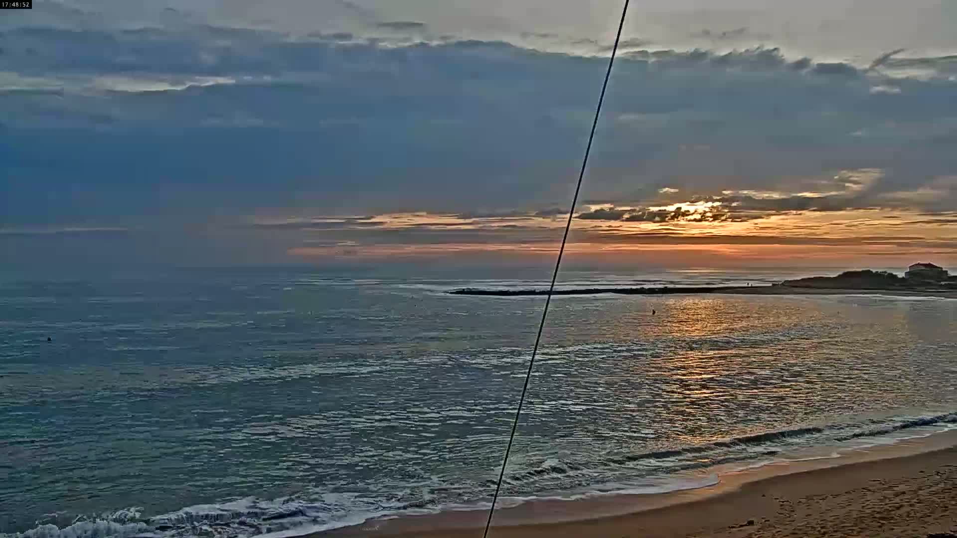 A sunrise casts orange and pink hues across a calm ocean with gentle waves lapping a sandy shore.