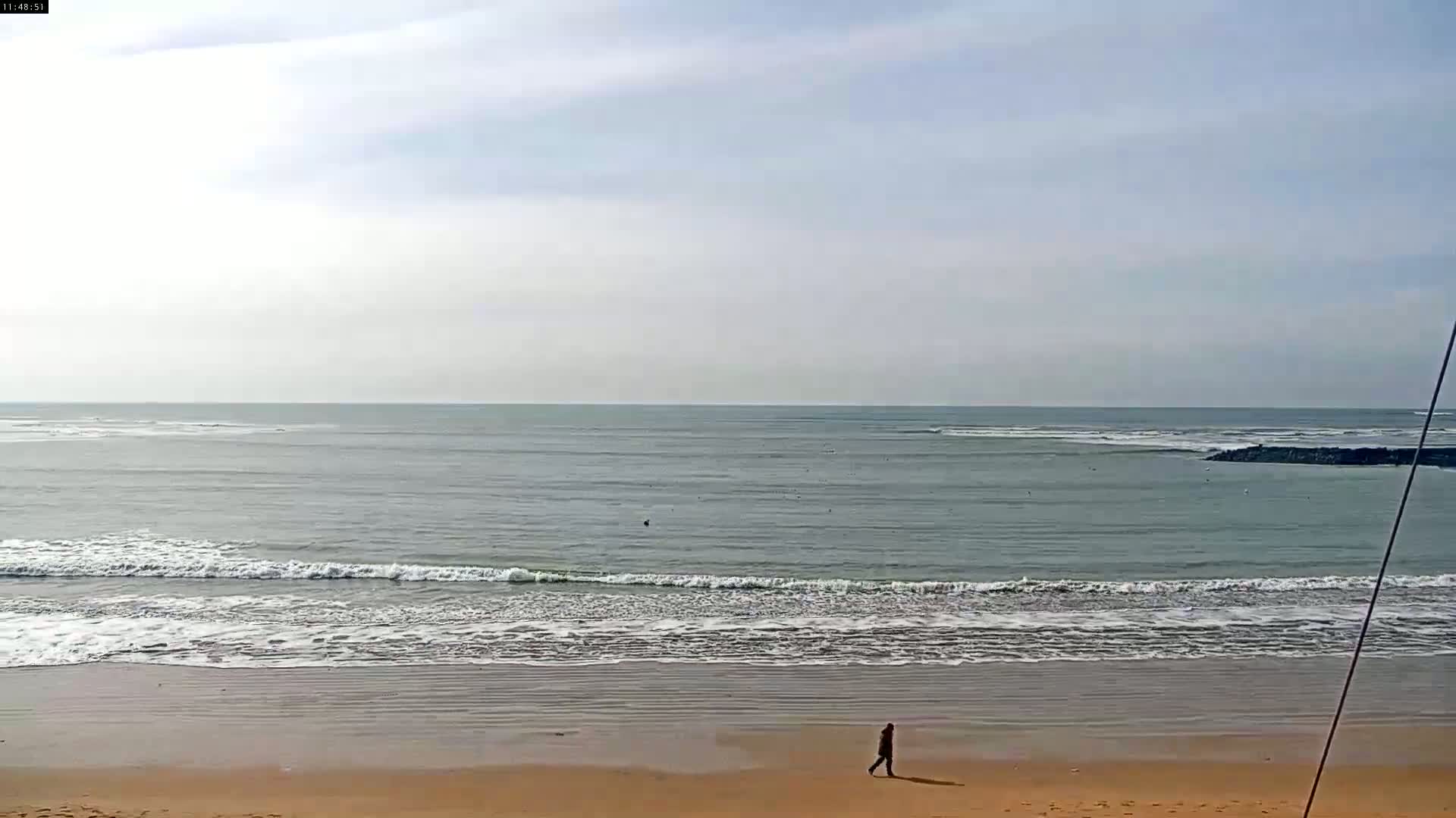 A person walks on a sandy beach near a calm ocean under a mostly cloudy sky.