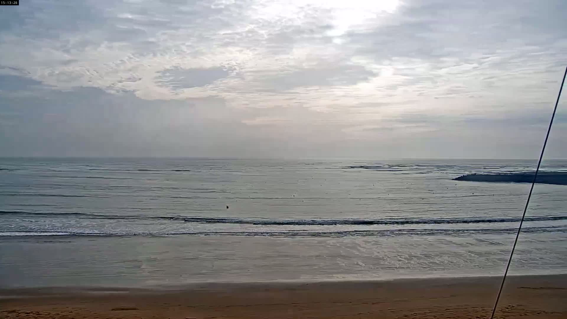 A sandy beach slopes down to a calm ocean with small waves under a mostly cloudy sky.