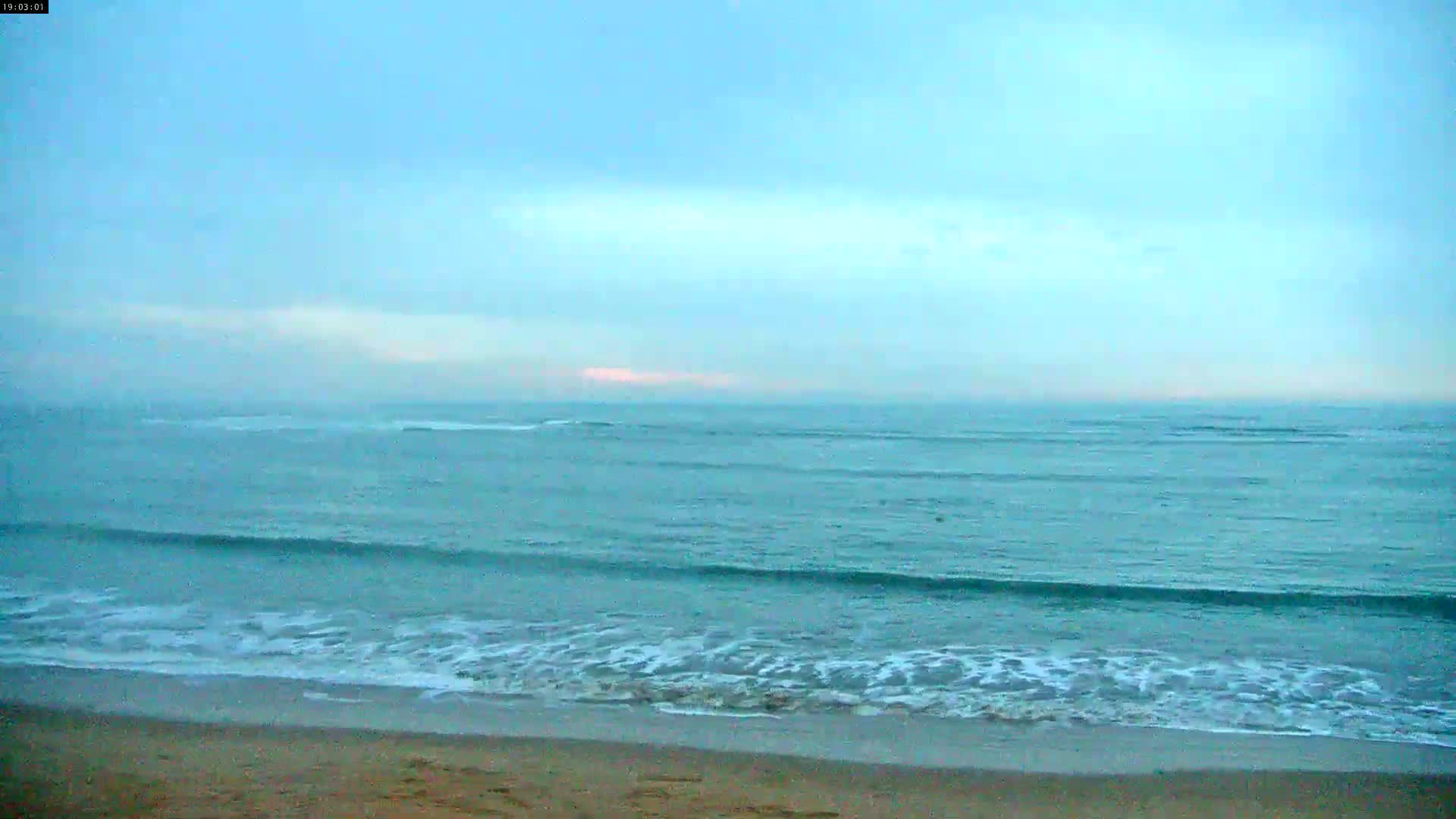 A sandy beach meets a calm, teal ocean under a pale blue, mostly clear sky.