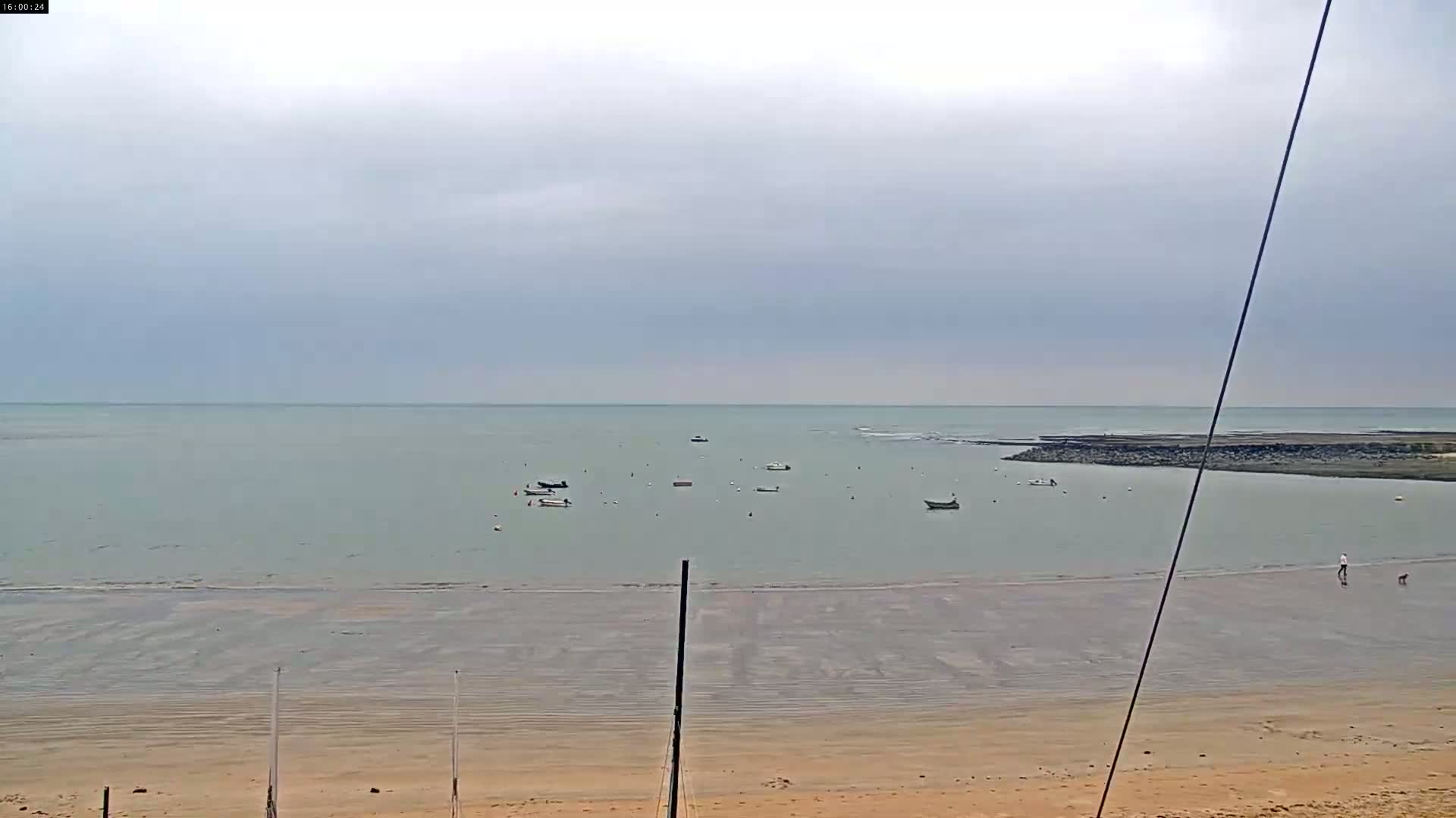 A calm, overcast day reveals a sandy beach with several small boats in the shallow water, a person walking a dog on the shore, and a rock jetty in the distance.