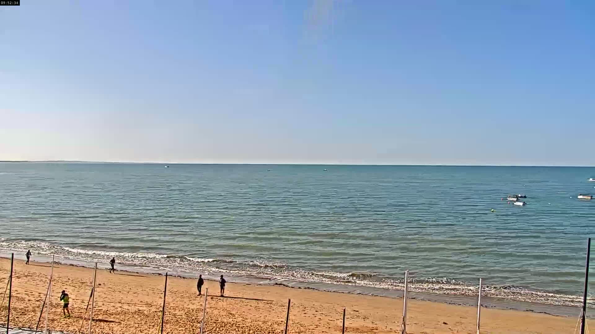 A sandy beach with several people walking along the shore meets a calm, blue ocean under a clear, sunny sky.
