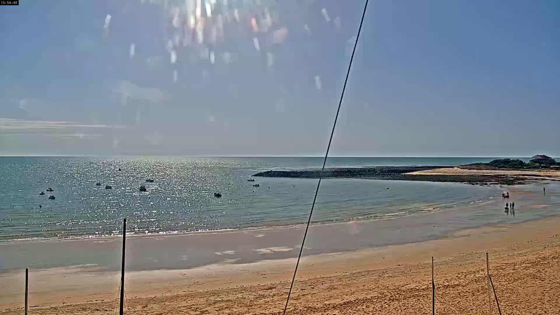 A sandy beach with calm, sparkling water extends to a rocky point, where several small boats are anchored under a sunny sky.