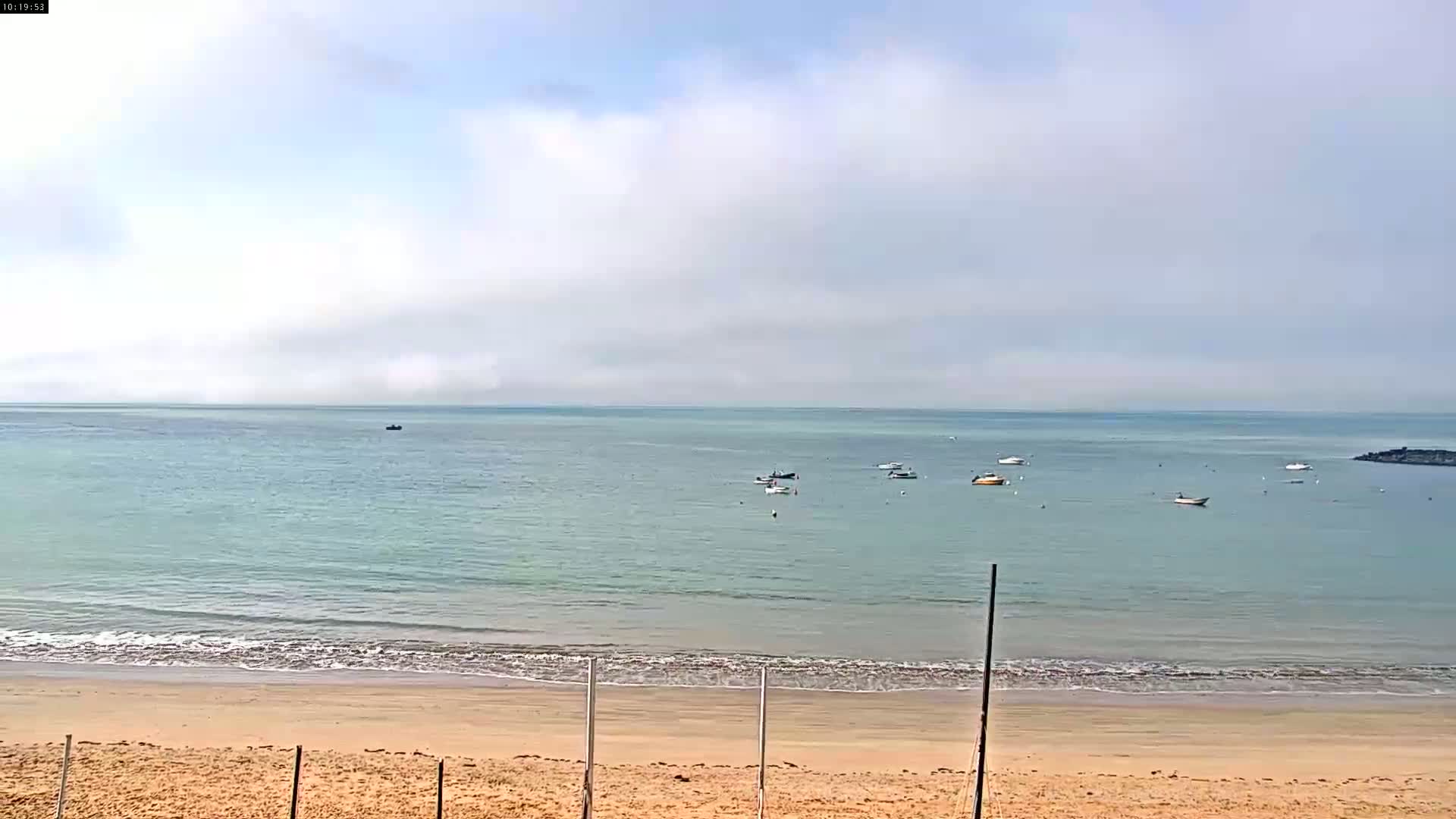 A sandy beach with calm, light-green water and several small boats visible in the distance under a partly cloudy sky.