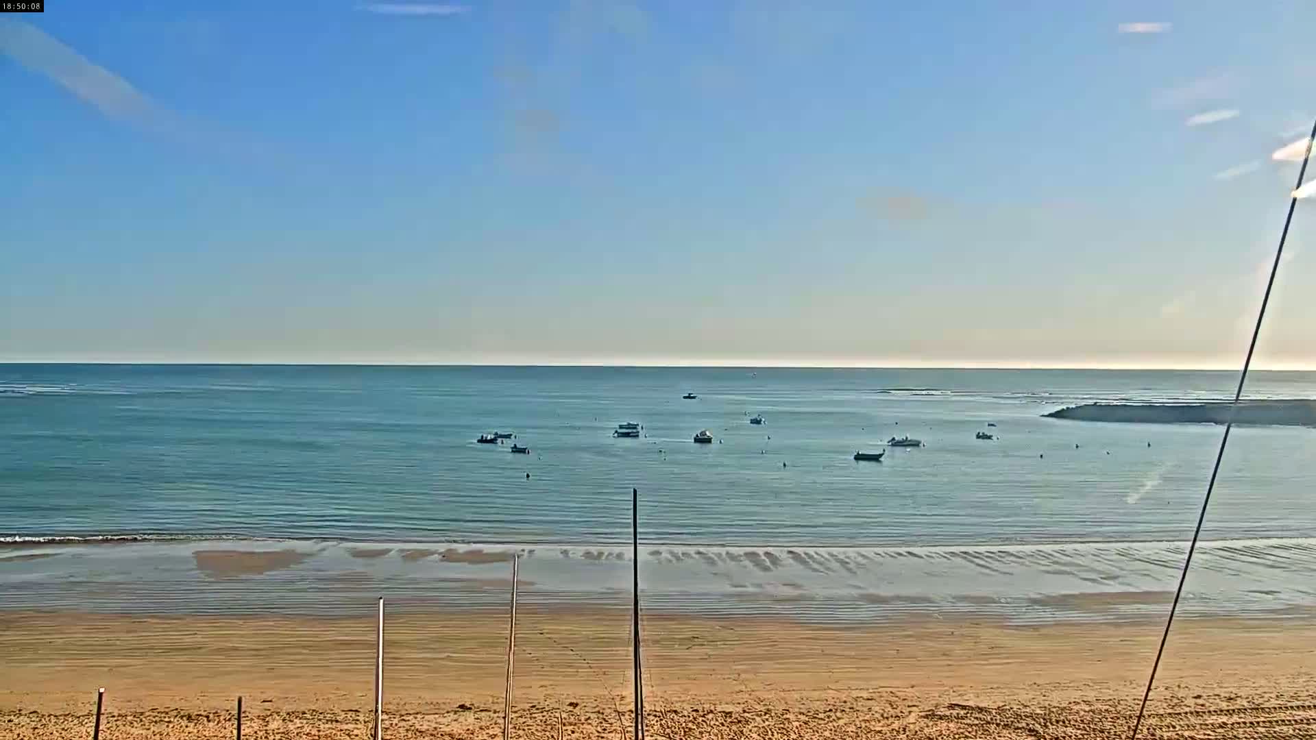 A sandy beach slopes down to a calm, blue ocean where several small boats are anchored; the sky is clear and bright.