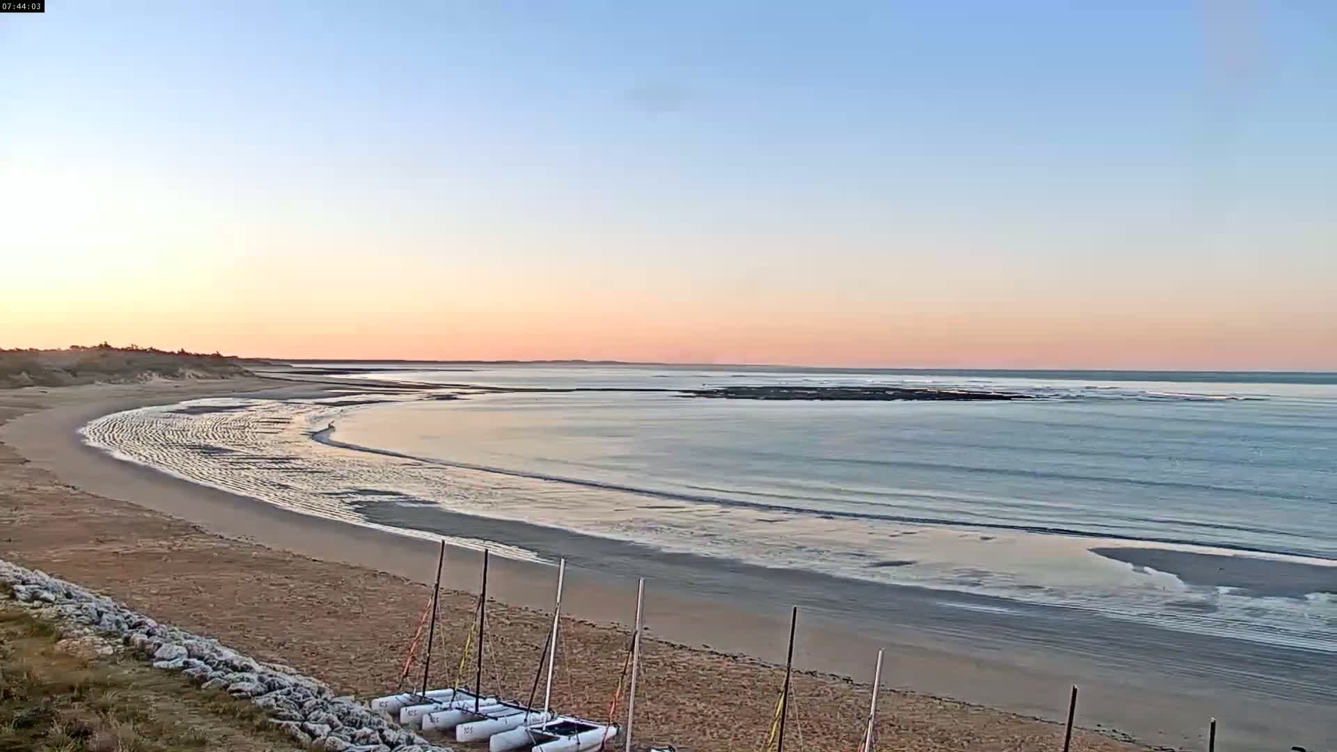 A sandy beach curves gently into a calm sea under a pale pink and blue sunset sky, with several small sailboats beached on the sand.