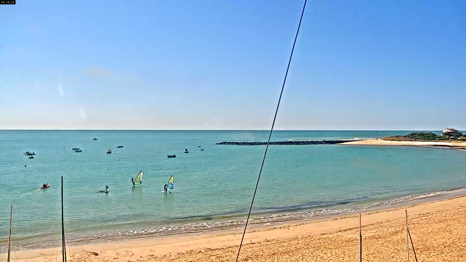 A sandy beach with calm, turquoise water features several sailboats and windsurfers on a sunny day.