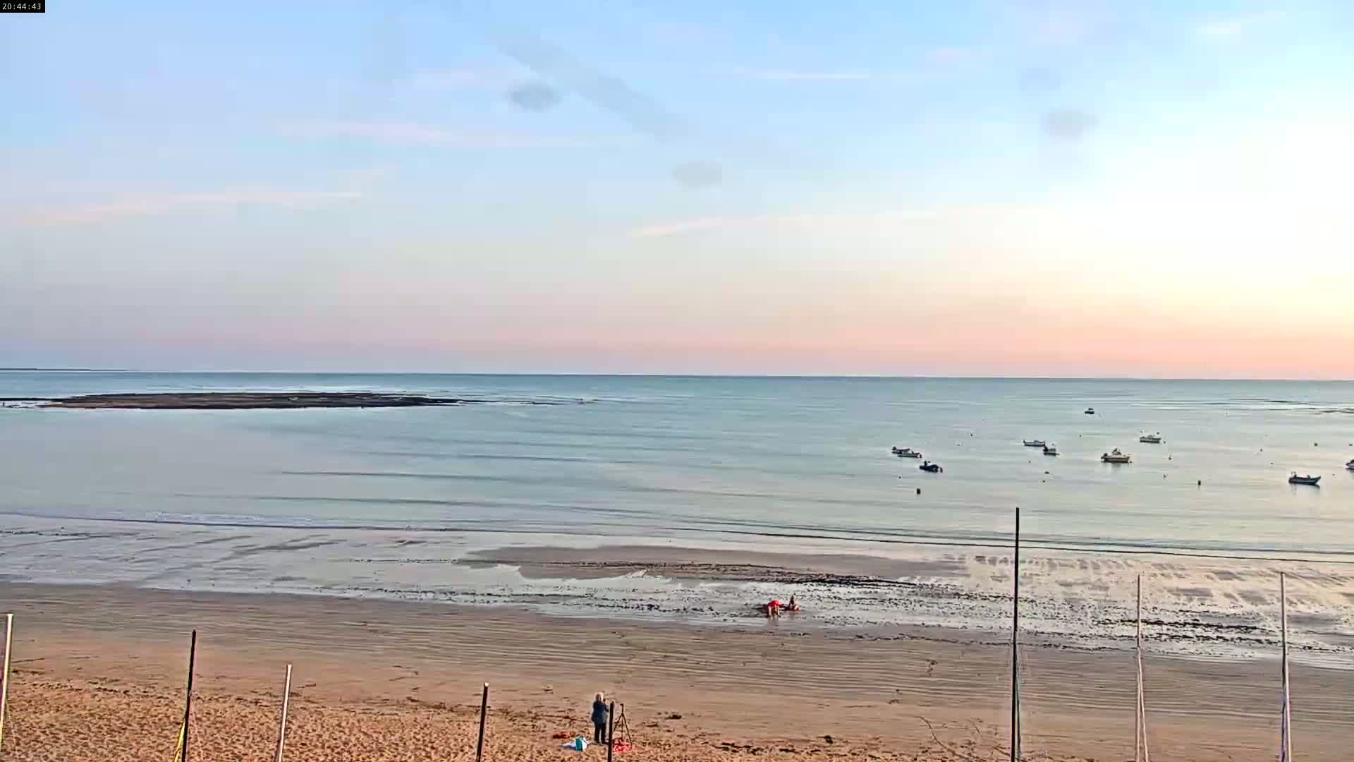 A calm, pale-pink sunset illuminates a sandy beach with several small boats in the distance and a few people on the shore.