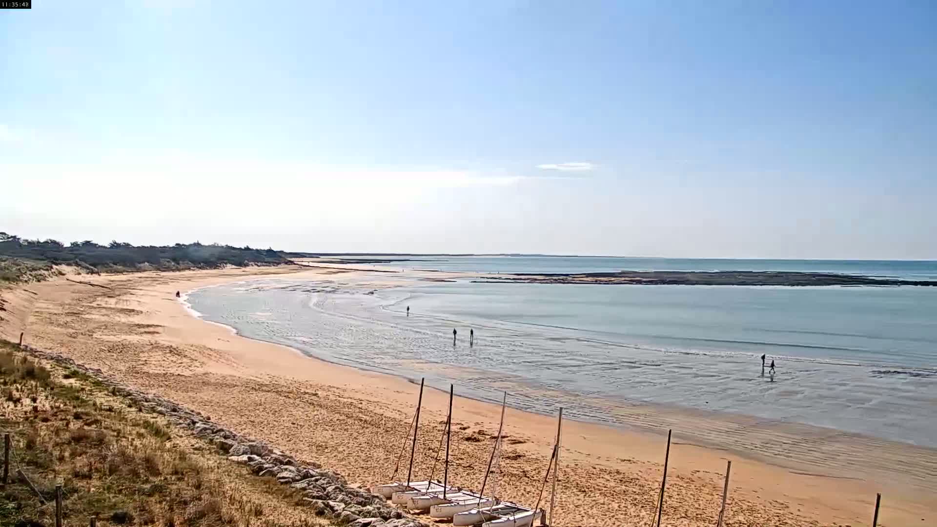 A sandy beach curves gently into a calm, light-blue ocean under a clear, sunny sky, with several small sailboats on the shore and a few people walking in the shallow water.
