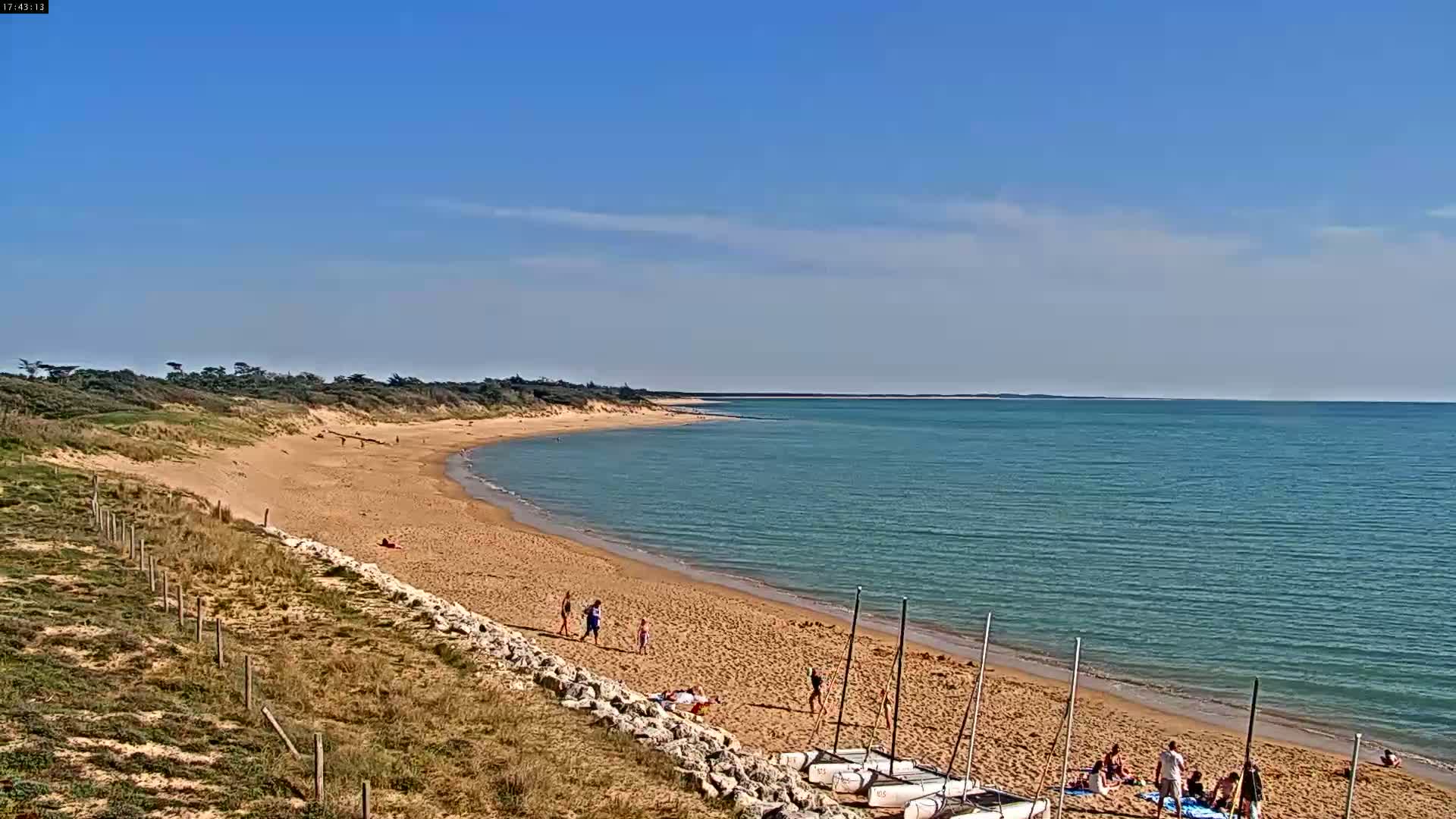 A sandy beach curves gently into a calm, teal sea under a mostly clear blue sky, with a few people enjoying the sunny day.