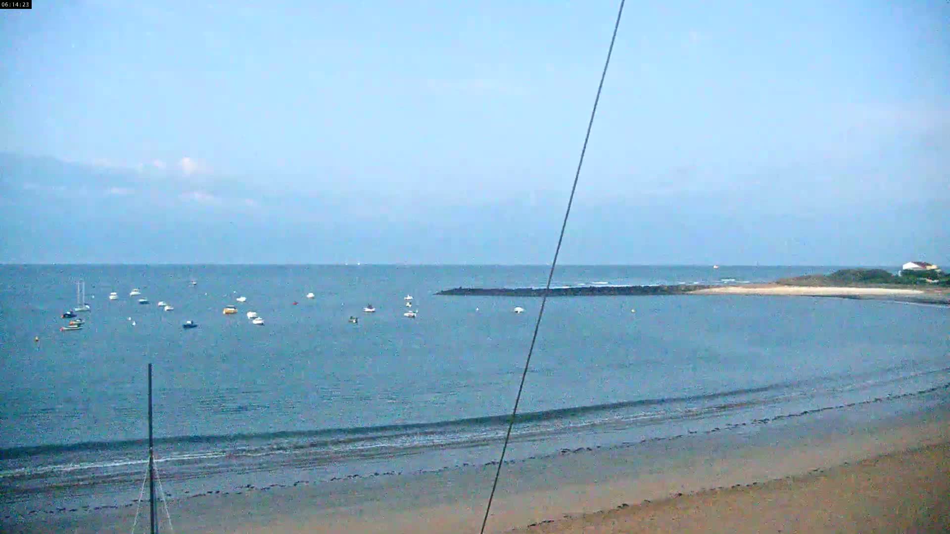 Several small boats are anchored in a calm bay under a clear, light-blue sky next to a sandy beach.