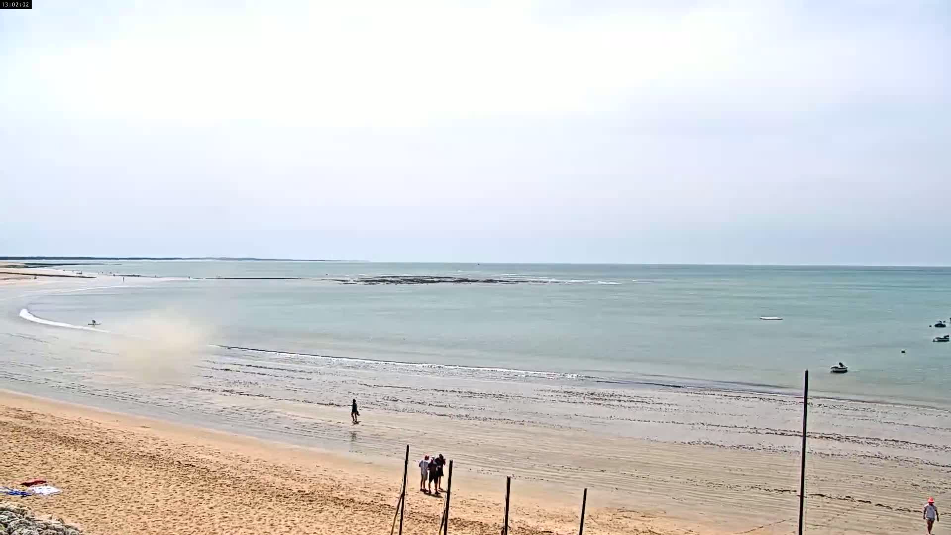 A sandy beach with a few people on it slopes gently down to a calm, light-green ocean under a mostly clear sky.