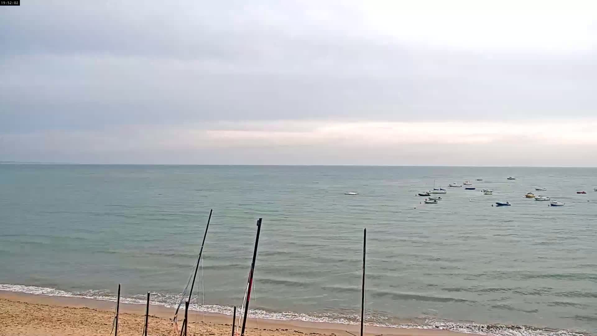 A calm, overcast day shows several small boats at anchor in a bay, with a sandy beach and tall posts in the foreground.