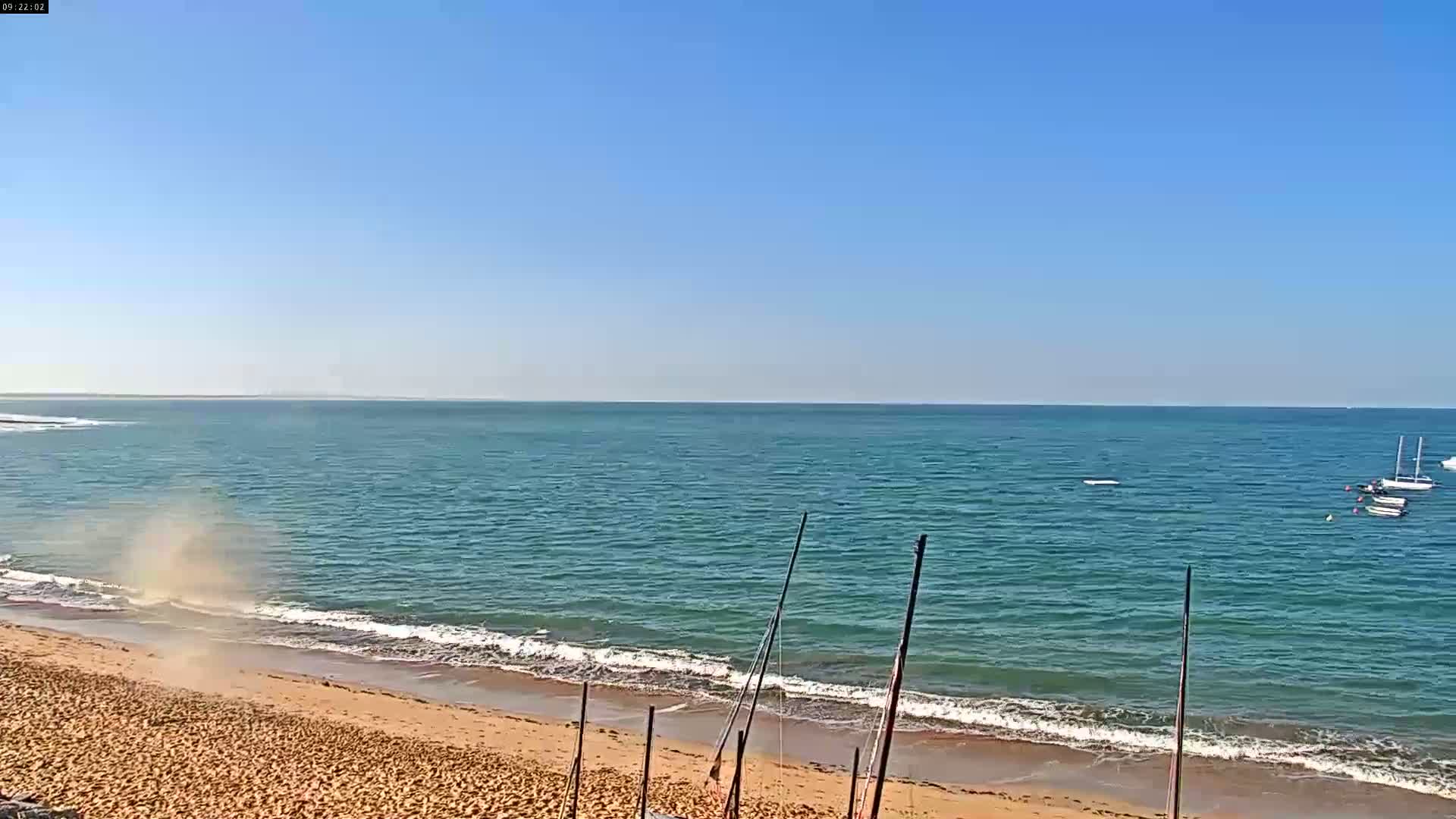 A sandy beach with small waves laps the shore of a calm, teal ocean under a clear blue sky; a small dust devil is visible near the shoreline.