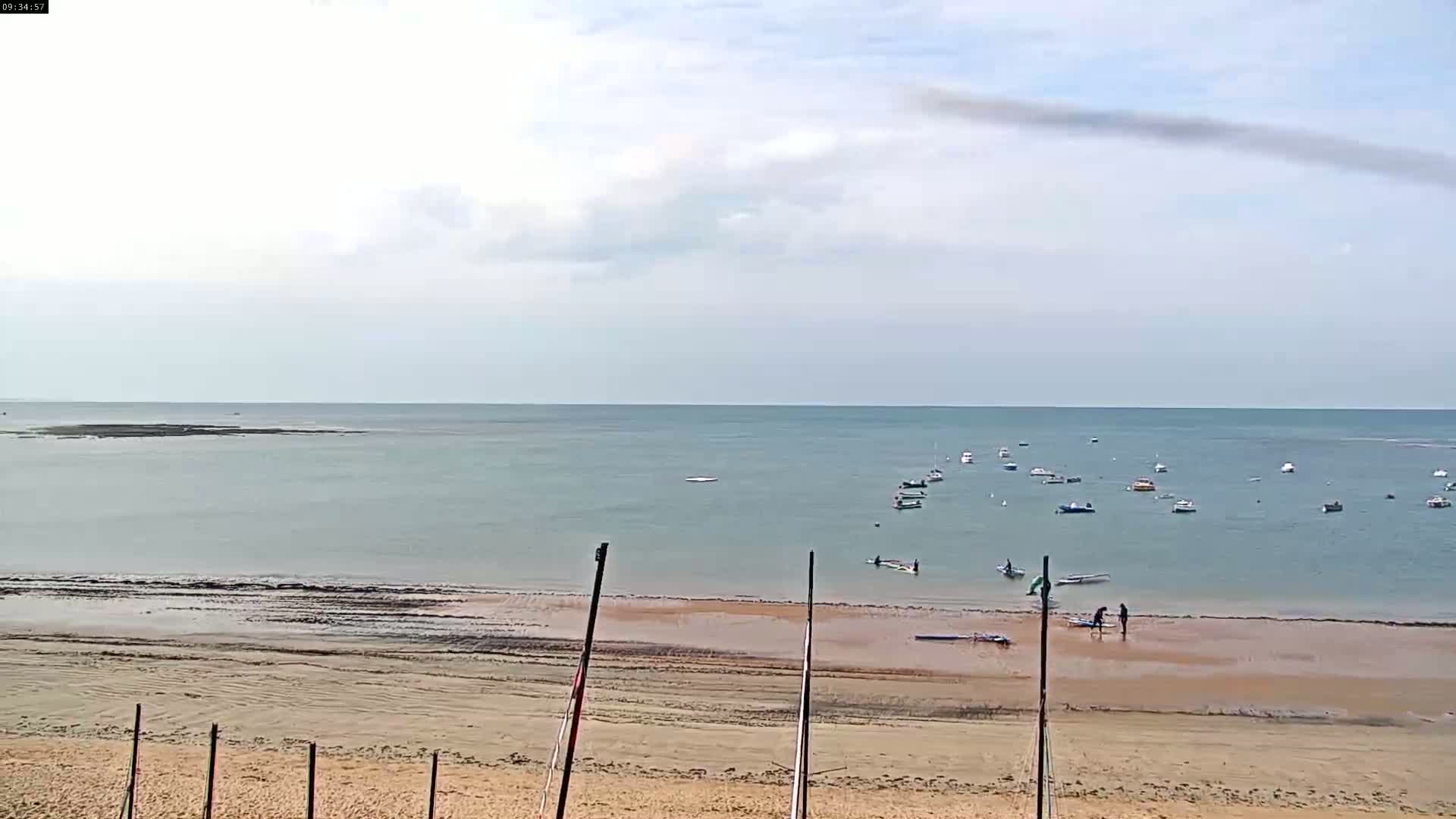 A sandy beach with several people near the water's edge, numerous small boats on calm, light-blue water under a mostly cloudy sky.