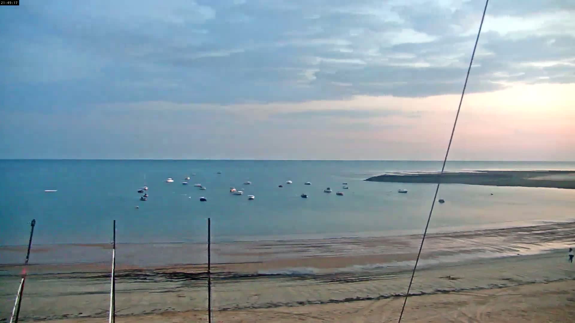 A calm, pale blue ocean with several small boats is visible from a sandy beach under a mostly cloudy sky.