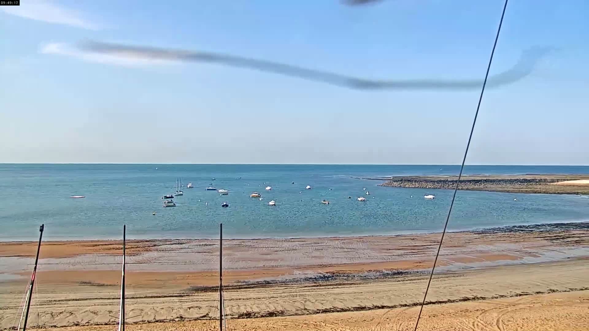 A sandy beach slopes down to a calm, blue bay where several small boats are anchored under a mostly clear sky.
