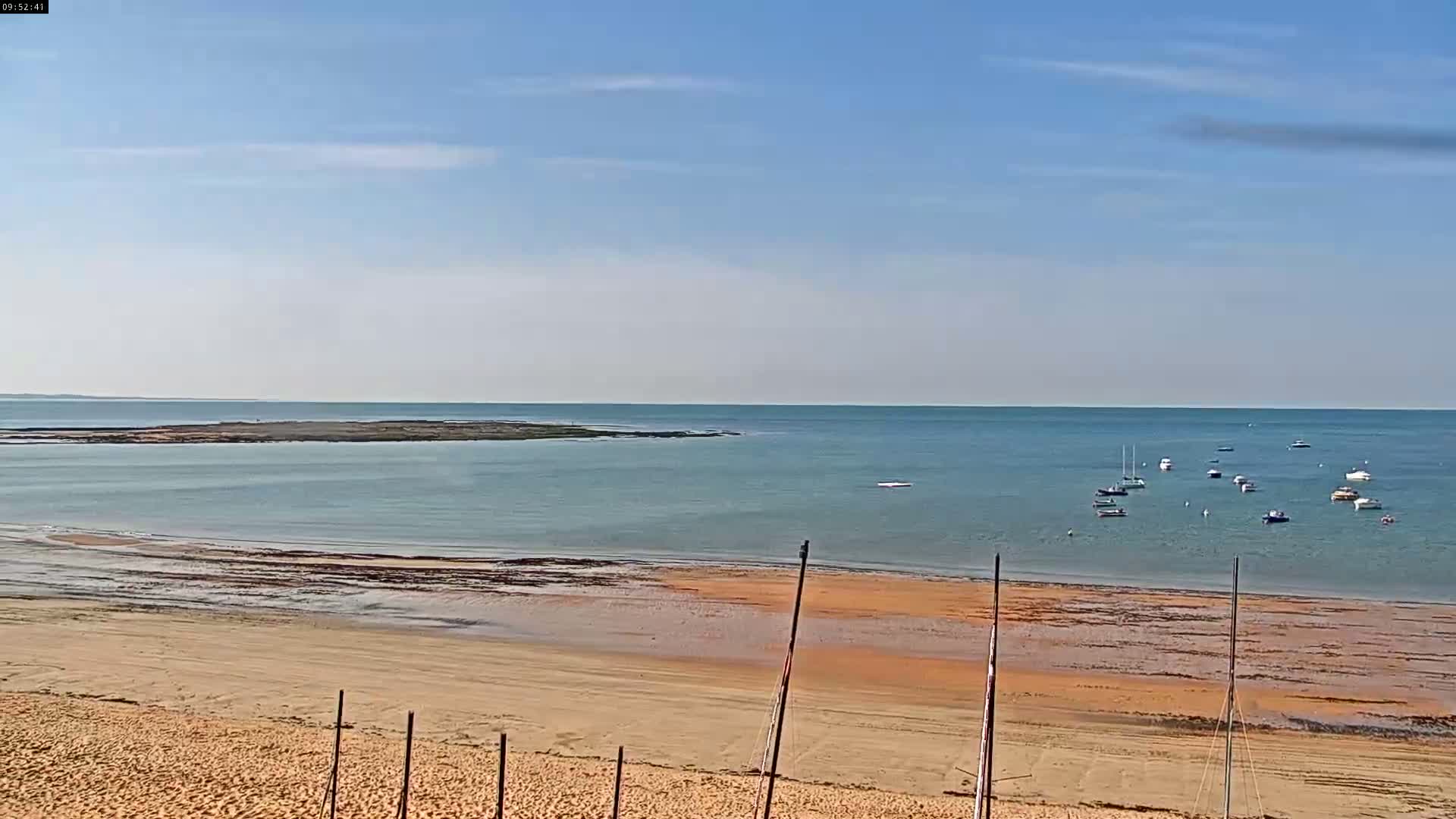 A sandy beach slopes down to a calm, light-blue ocean where several small boats are anchored under a mostly clear, light-blue sky.