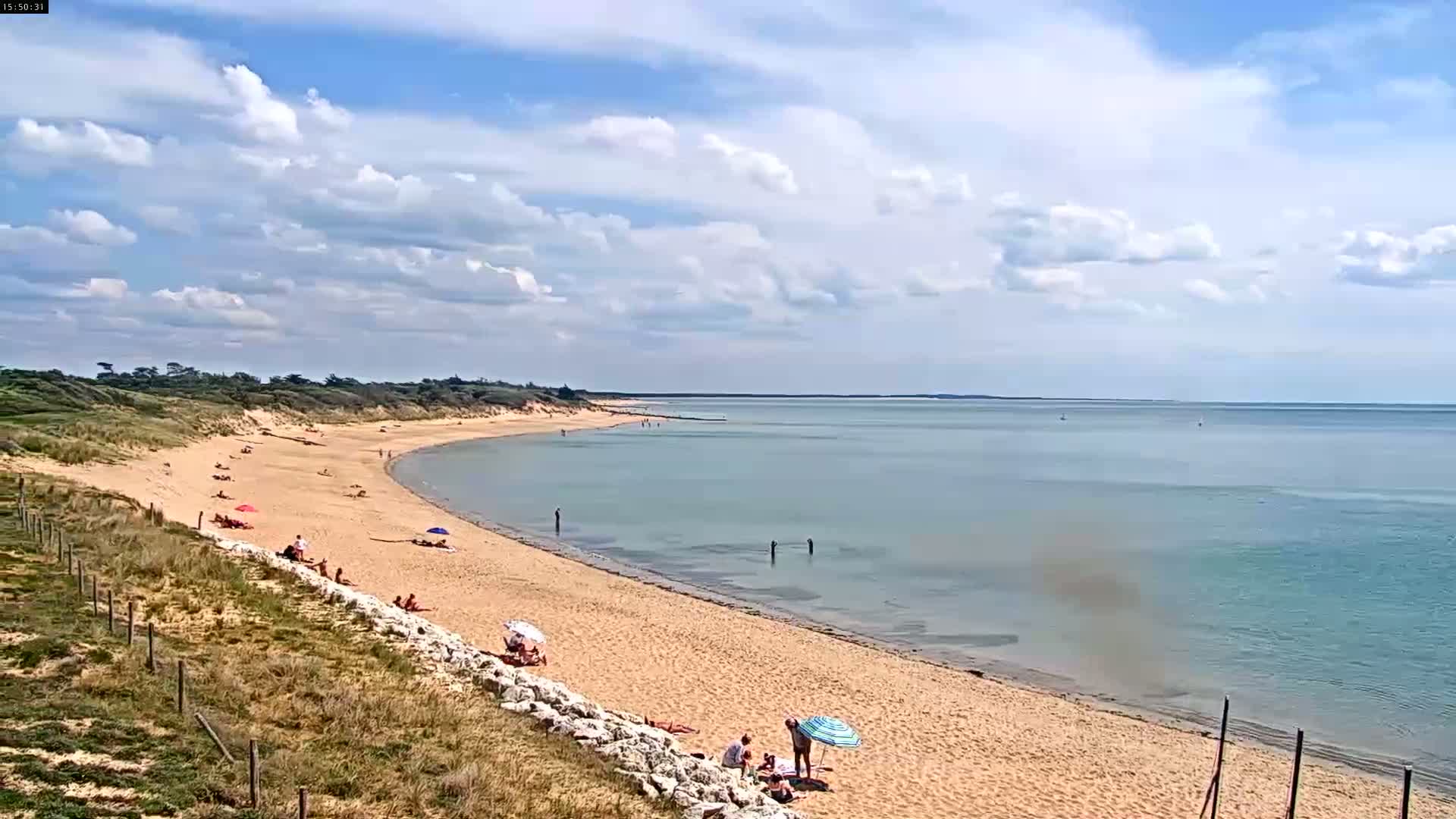 A sandy beach curves gently into a calm, light-blue sea under a partly cloudy sky.