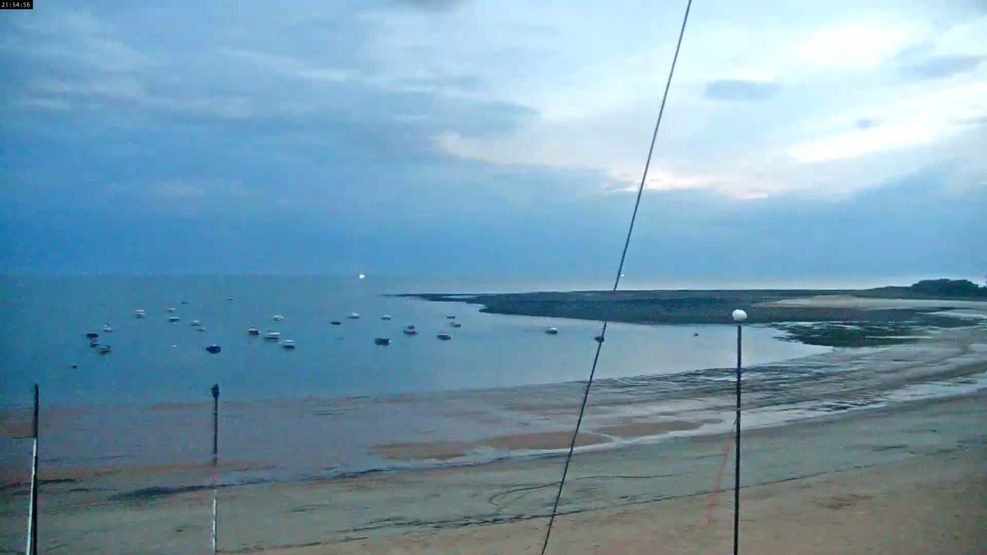 A mostly cloudy, dusky sky overlooks a calm bay with several small boats anchored near a sandy beach at low tide.