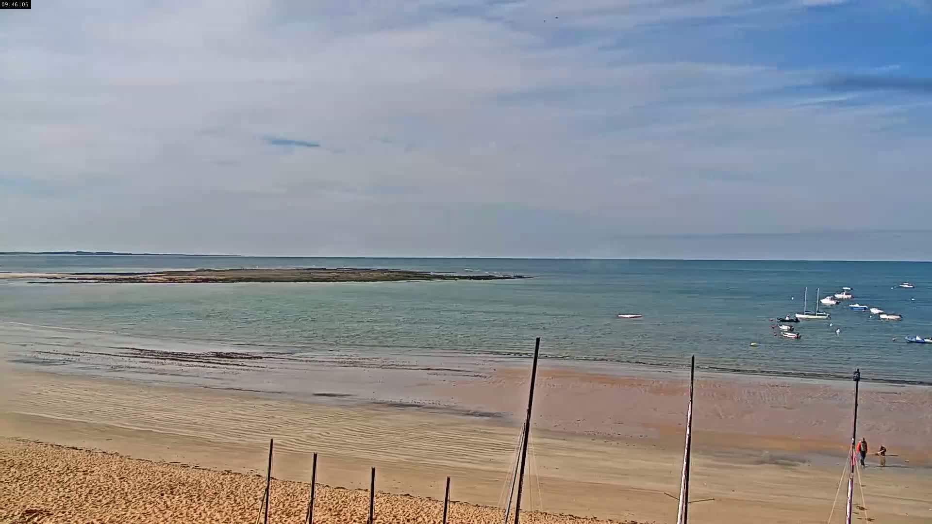 A sandy beach stretches to a calm, light-green ocean where several small boats are anchored near a small, dark island under a partly cloudy sky.