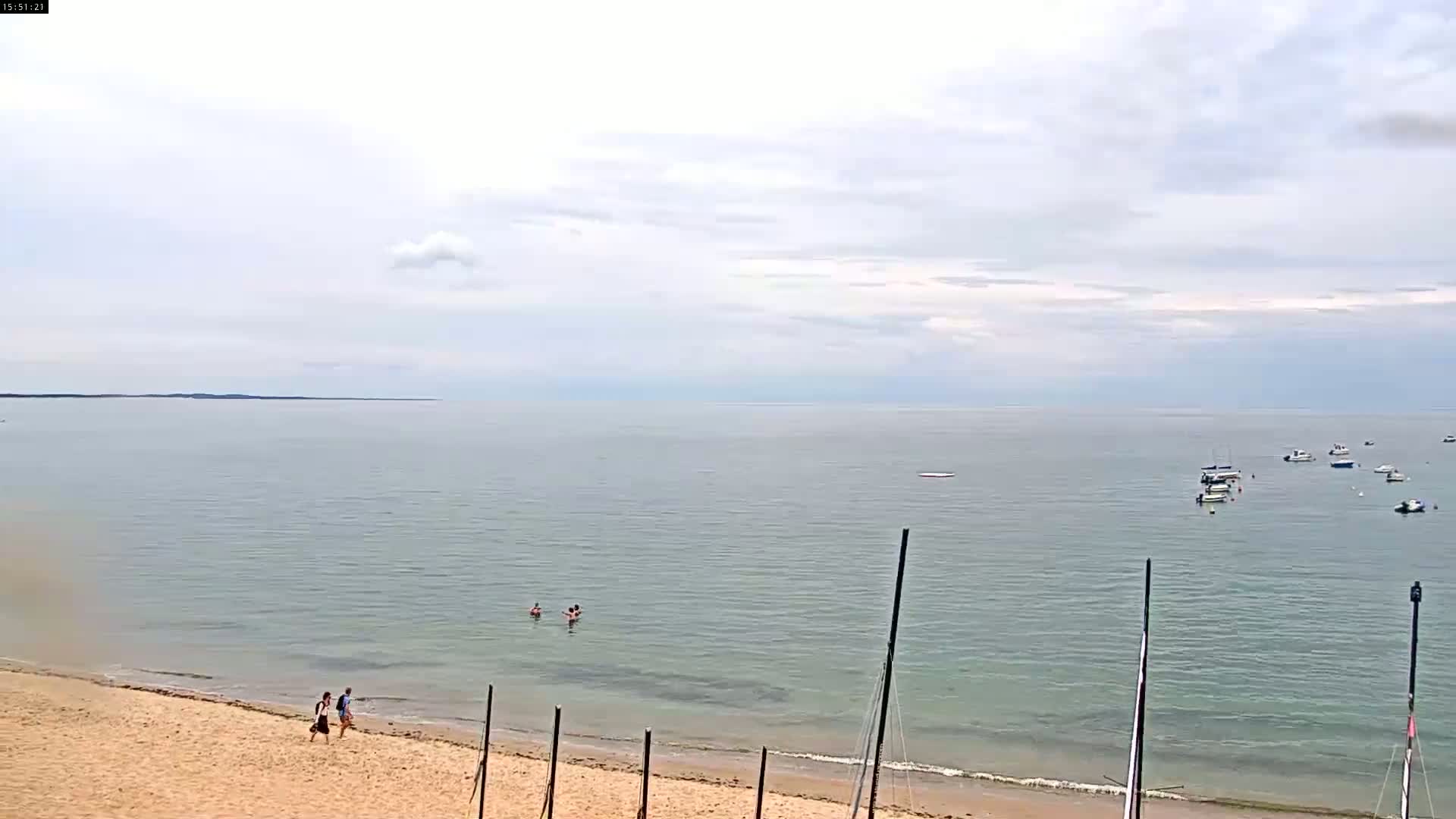 A sandy beach with two people walking along the shore, several people swimming in calm, grey water, and a few boats in the distance under a cloudy sky.