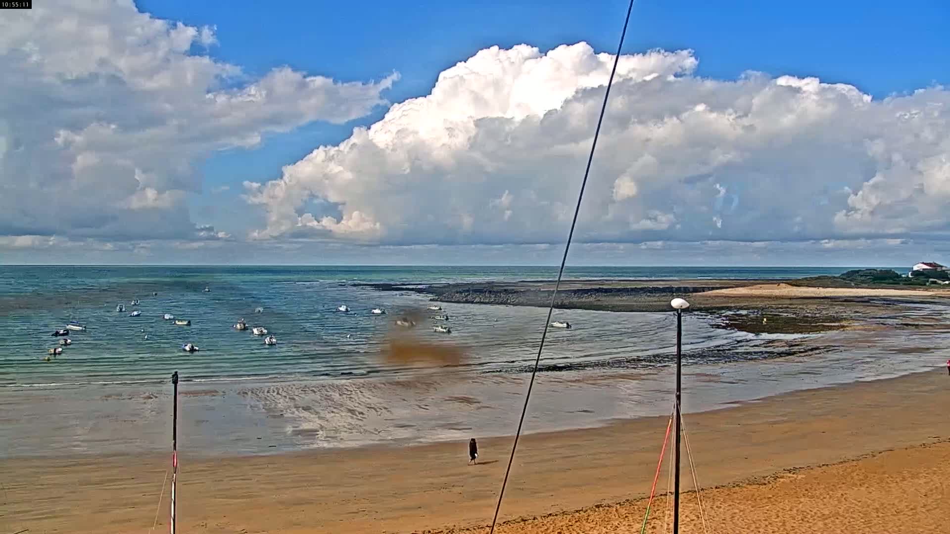 A sandy beach stretches out to a calm sea where numerous small boats are anchored under a partly cloudy sky.