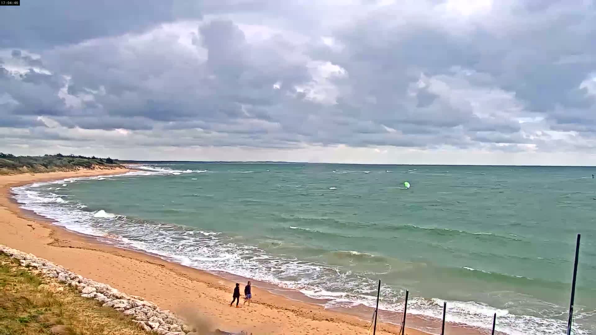 Two people walk along a sandy beach with small waves lapping the shore under a cloudy sky, with windsurfers visible in the distance on the water.