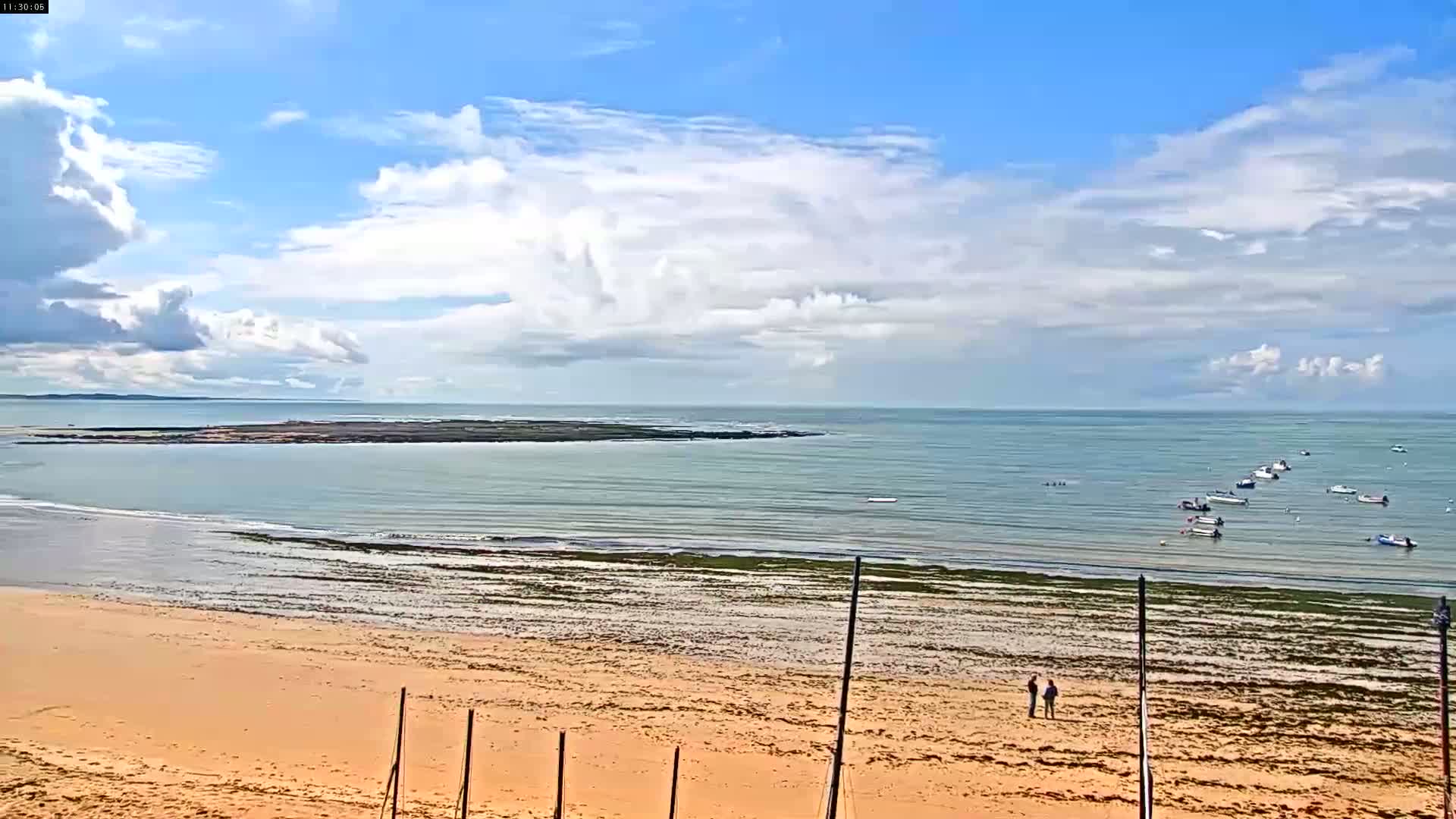 A sandy beach stretches to a calm sea dotted with several small boats under a partly cloudy sky.