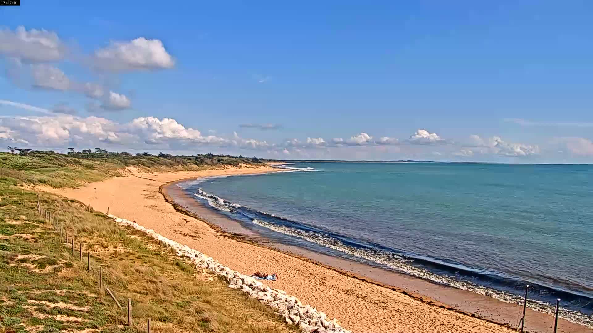 A sandy beach curves gently into a calm, teal ocean under a partly cloudy blue sky.