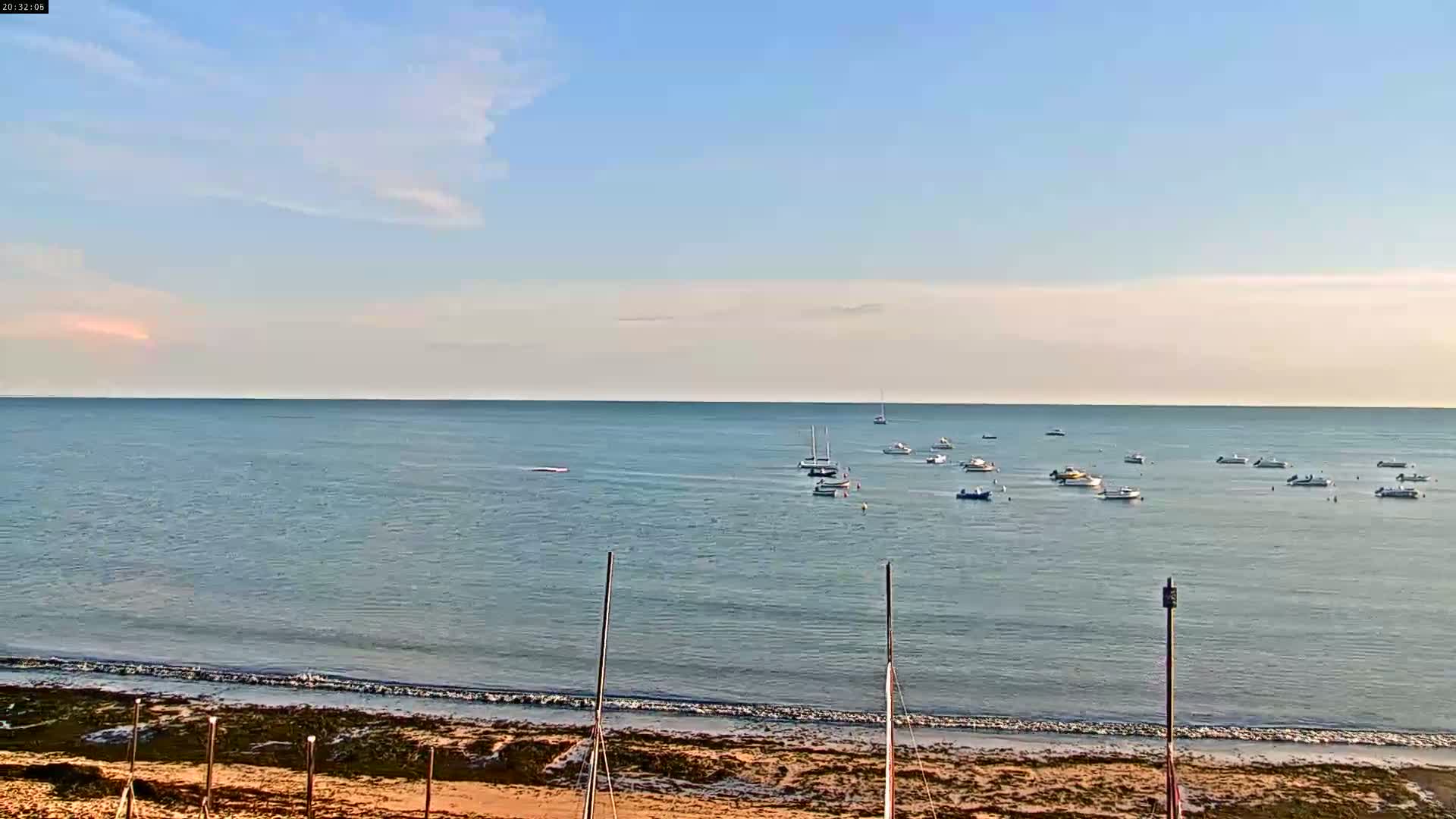 A calm, mostly clear day at the beach shows numerous small boats anchored in the bay, with a sandy shore and a few poles in the foreground.